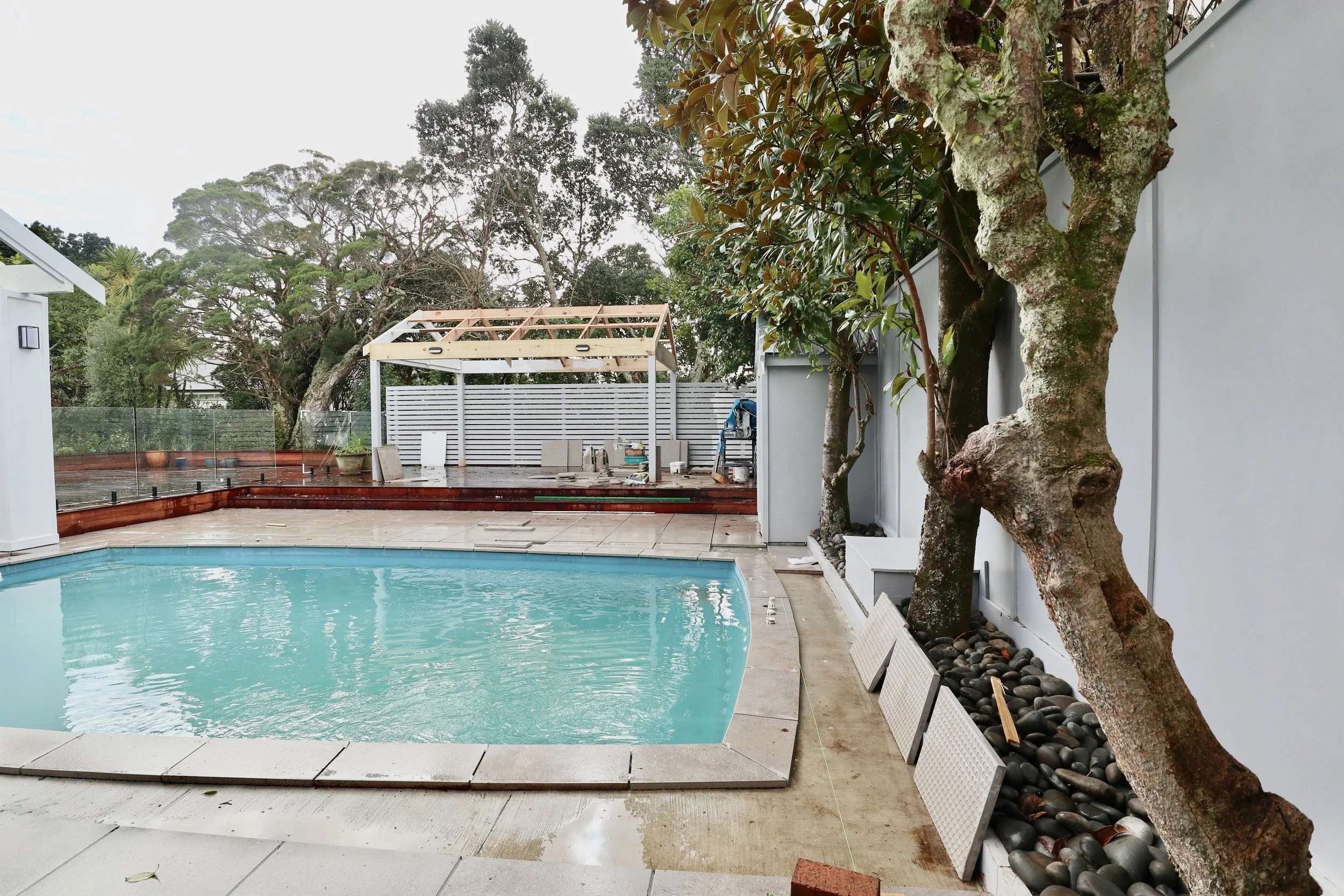 A backyard swimming pool with a patio area, trees along the side, and a white privacy fence. The patio area has construction materials and an unfinished wooden pergola frame.