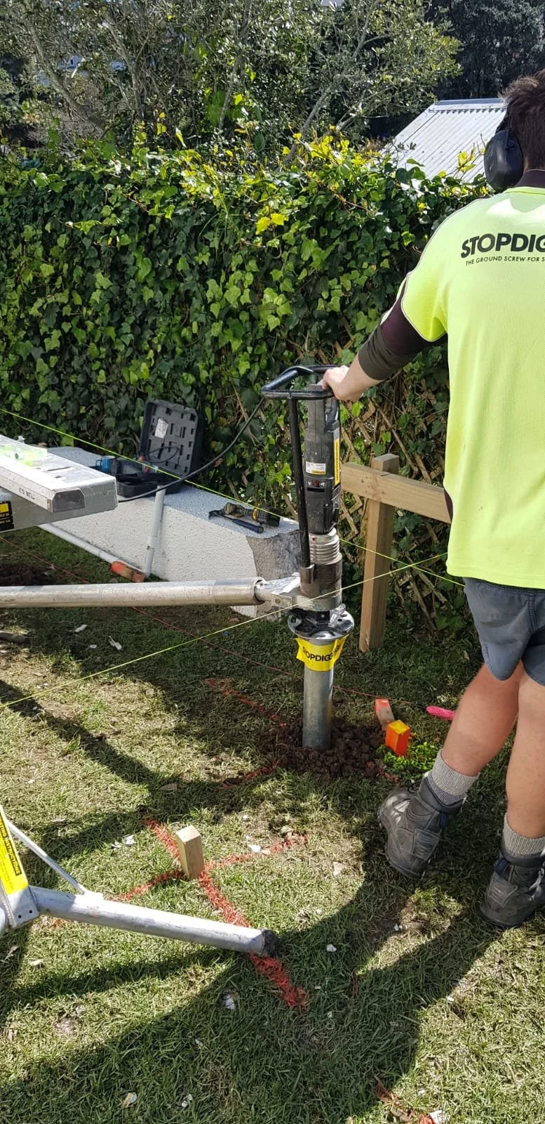 Person operating a ground screw installation machine with a bright yellow safety vest and headphones, set up on grass with orange spray paint markings, next to a hedge and a workbench with tools and a case.