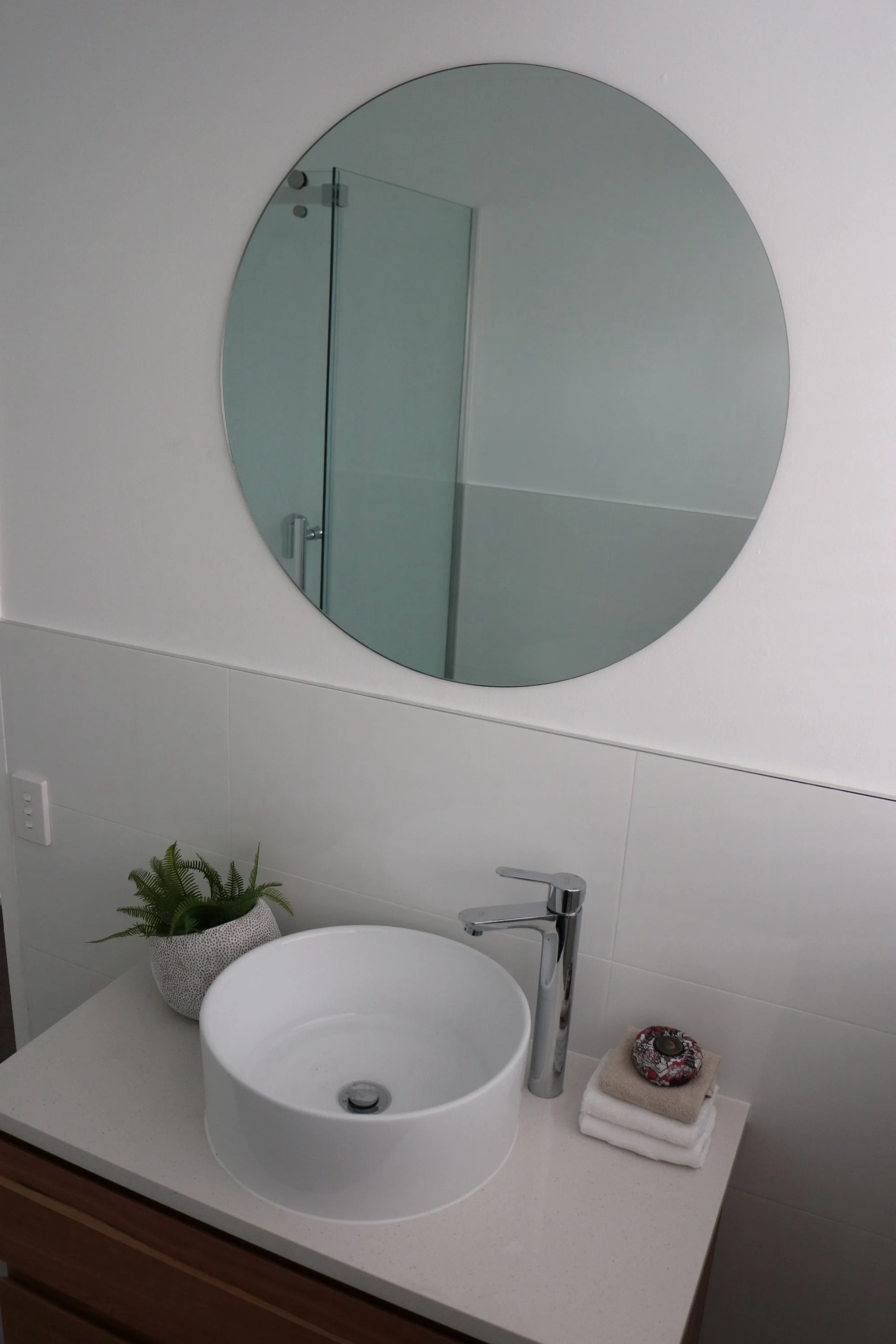 A bathroom vanity with a round white vessel sink, a modern chrome faucet, a small potted fern, a stack of white towels, and a decorative soap dish. A round mirror is mounted on the wall above the sink, and a glass shower door is reflected in the mirr