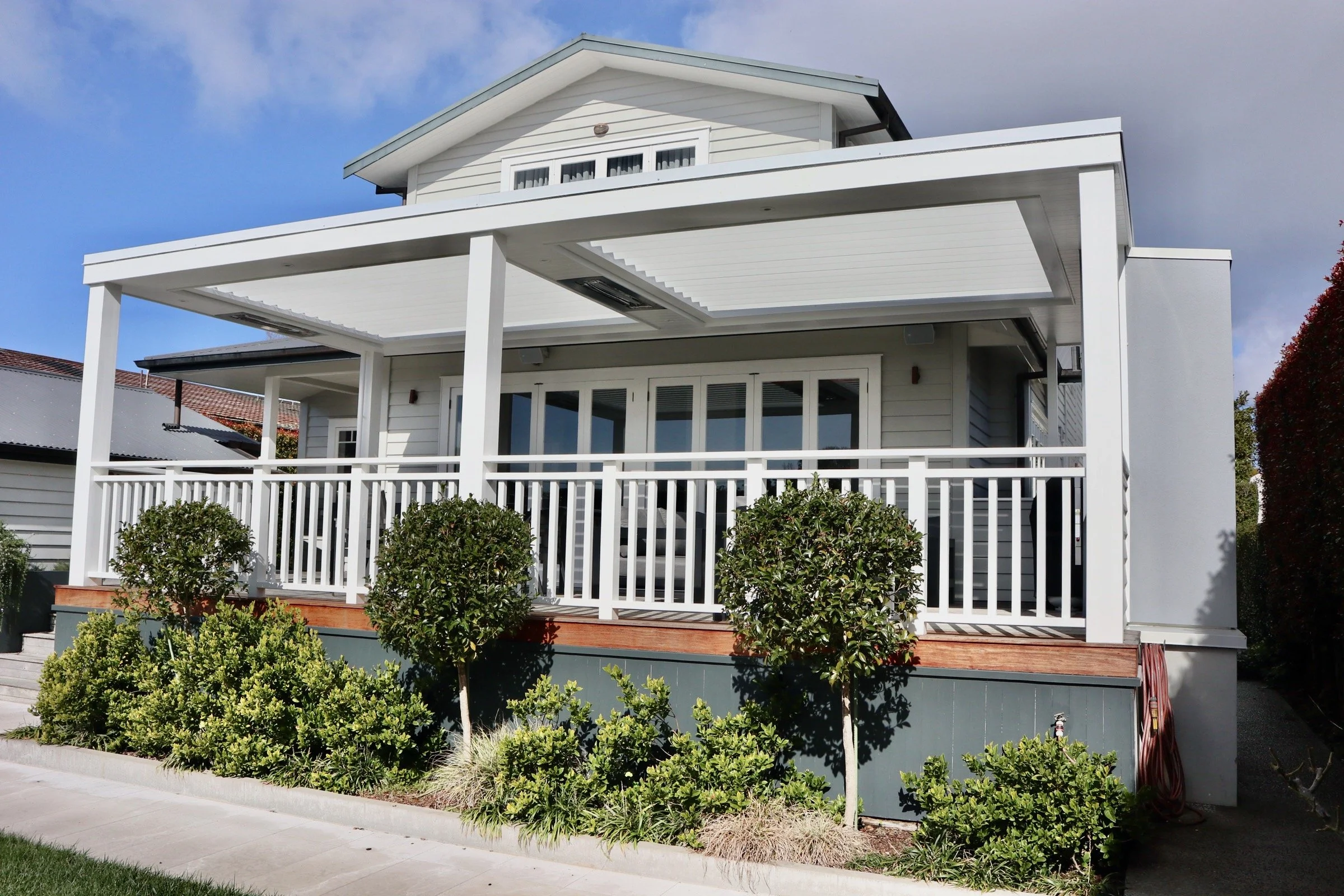 White two-story house with a porch, white railing, and surrounding shrubbery under a partly cloudy sky.