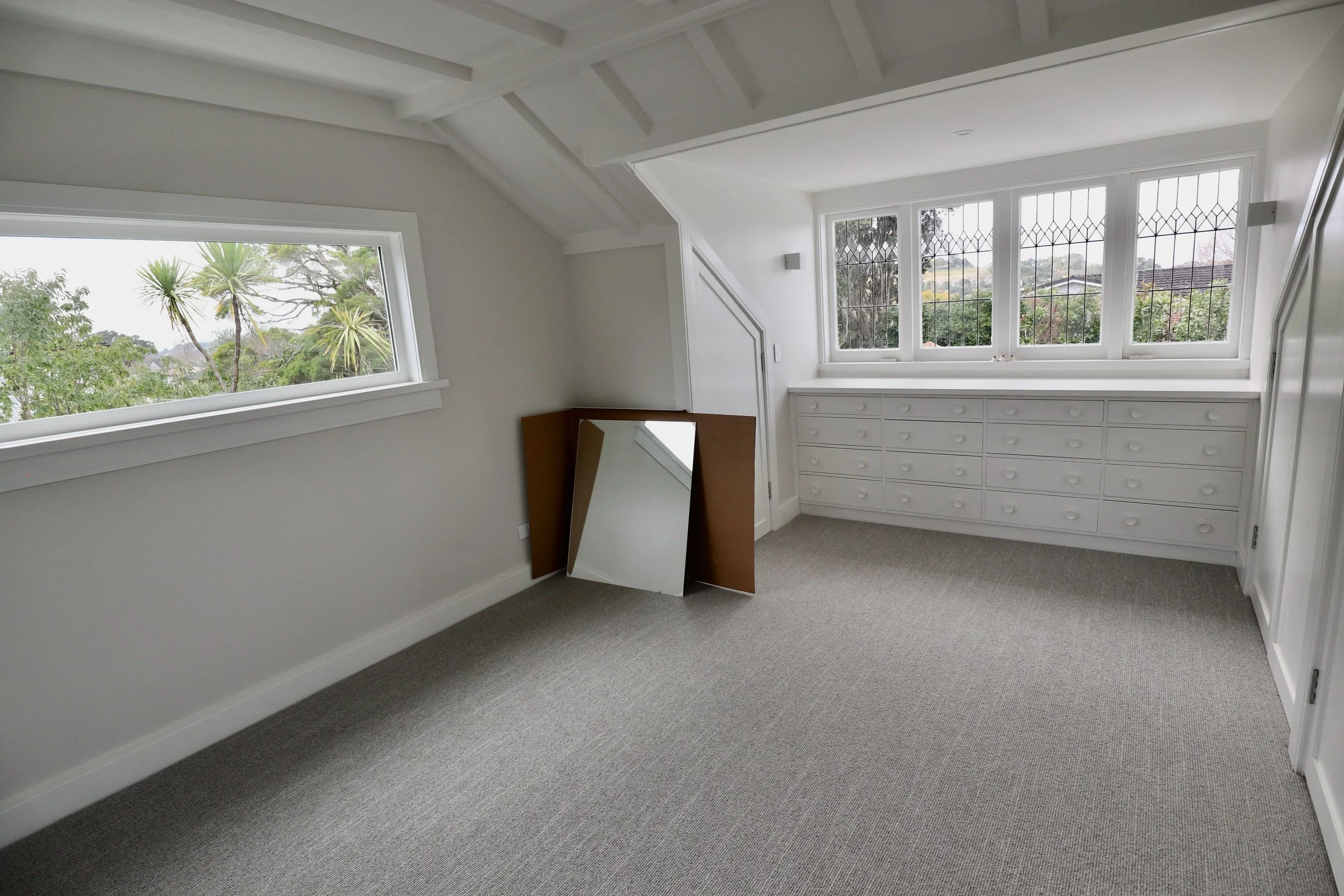 Empty room with large windows, white walls, gray carpet, and a leaning mirror.