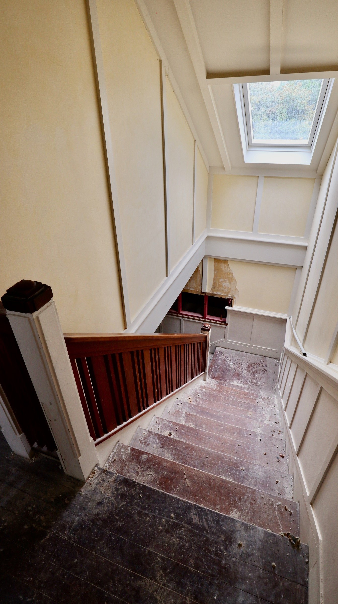 Looking down a staircase under renovation with a window at the top, painted yellow walls, a wooden handrail, and dust on the steps.