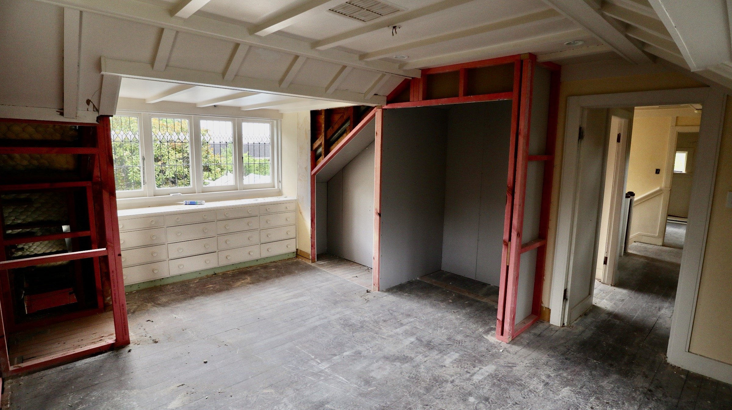 Room under construction with exposed framing, a large window with diamond-patterned bars, and a set of small white drawers beneath the window.