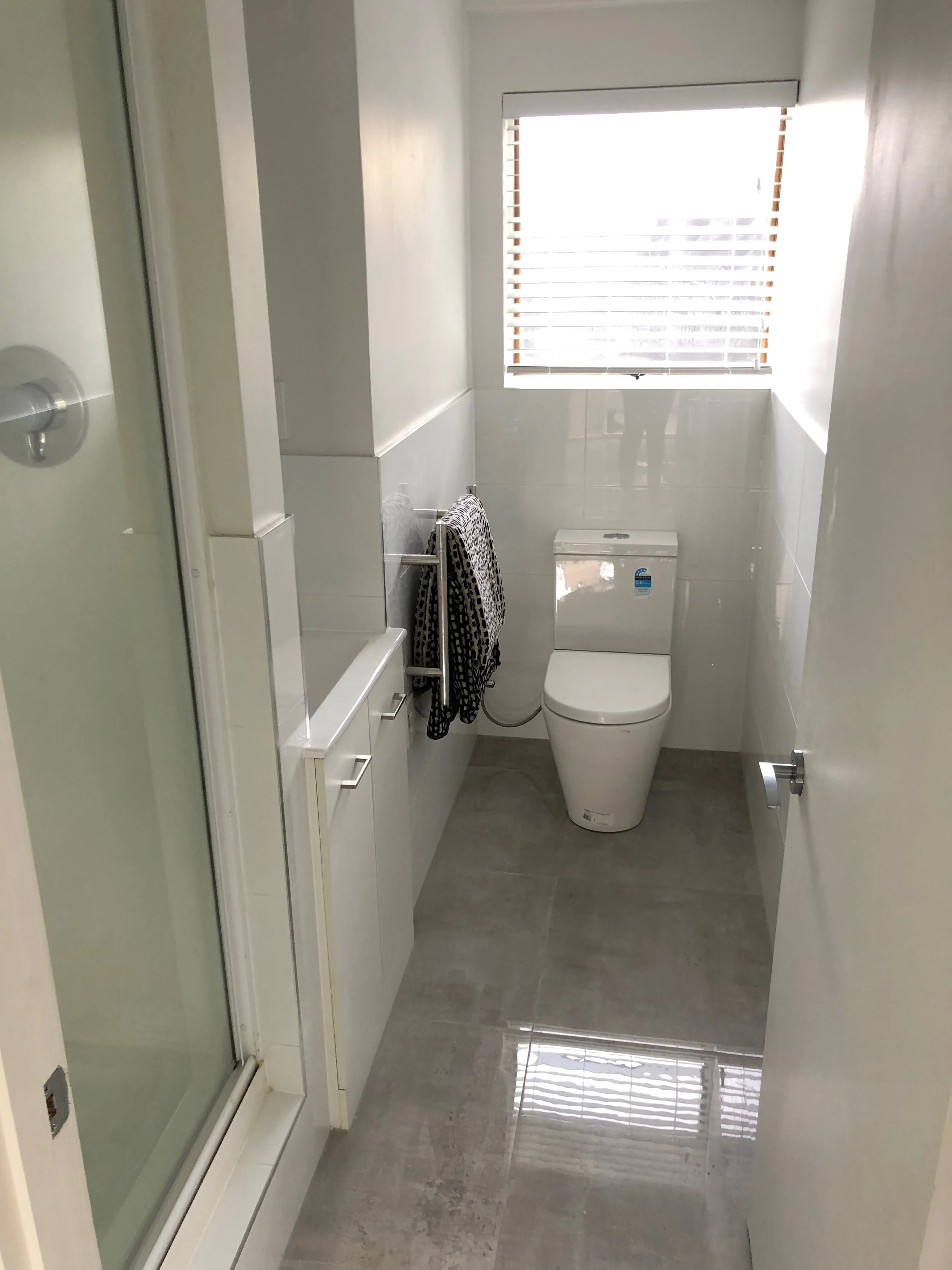 Small modern bathroom with a toilet, window with blinds, and a shower stall, all in white and gray tones.