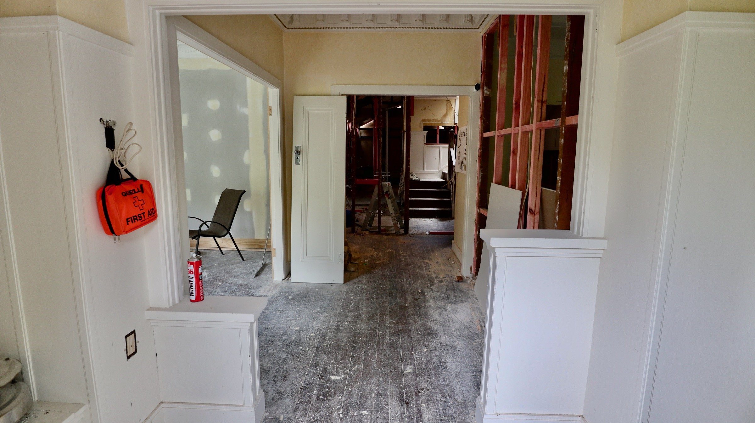 Room under renovation with exposed wooden framing on the right and white wainscoting on the left. A red first aid bag and a fire extinguisher are mounted on the left wall. The floor is unfinished and covered in dust. In the background, there are stai