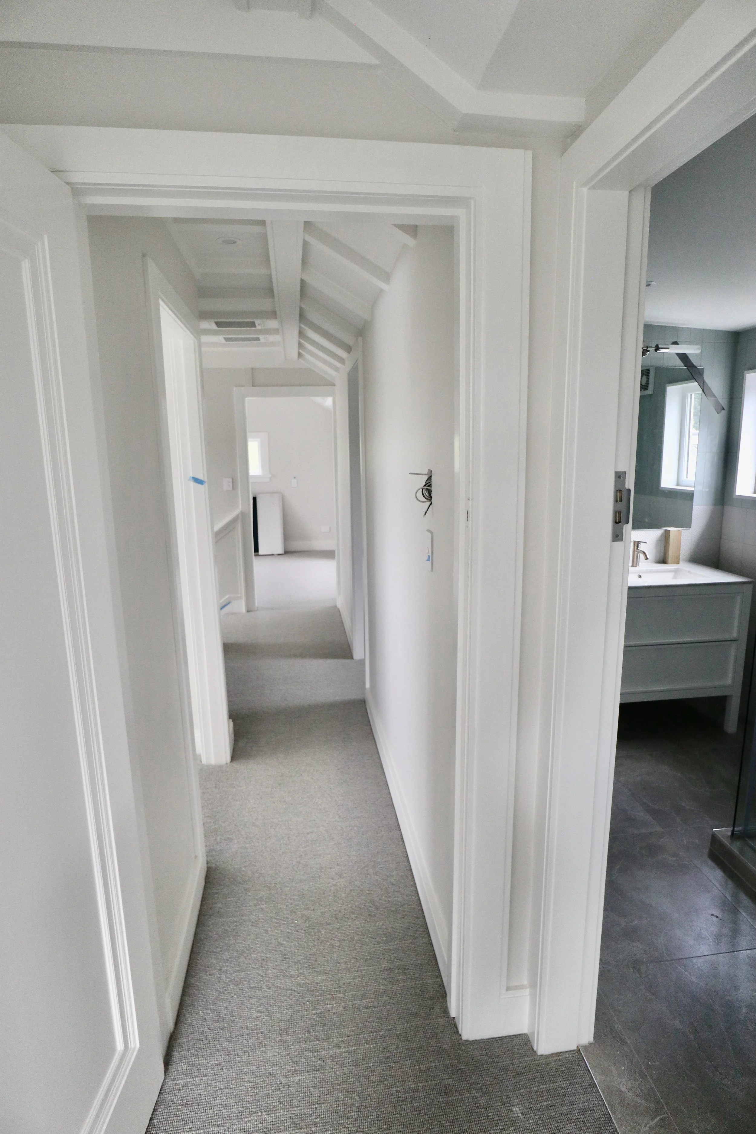A bright hallway in a modern house with white walls, carpeted floor, and visible doorways to other rooms, including a bathroom with gray tiles and a vanity.