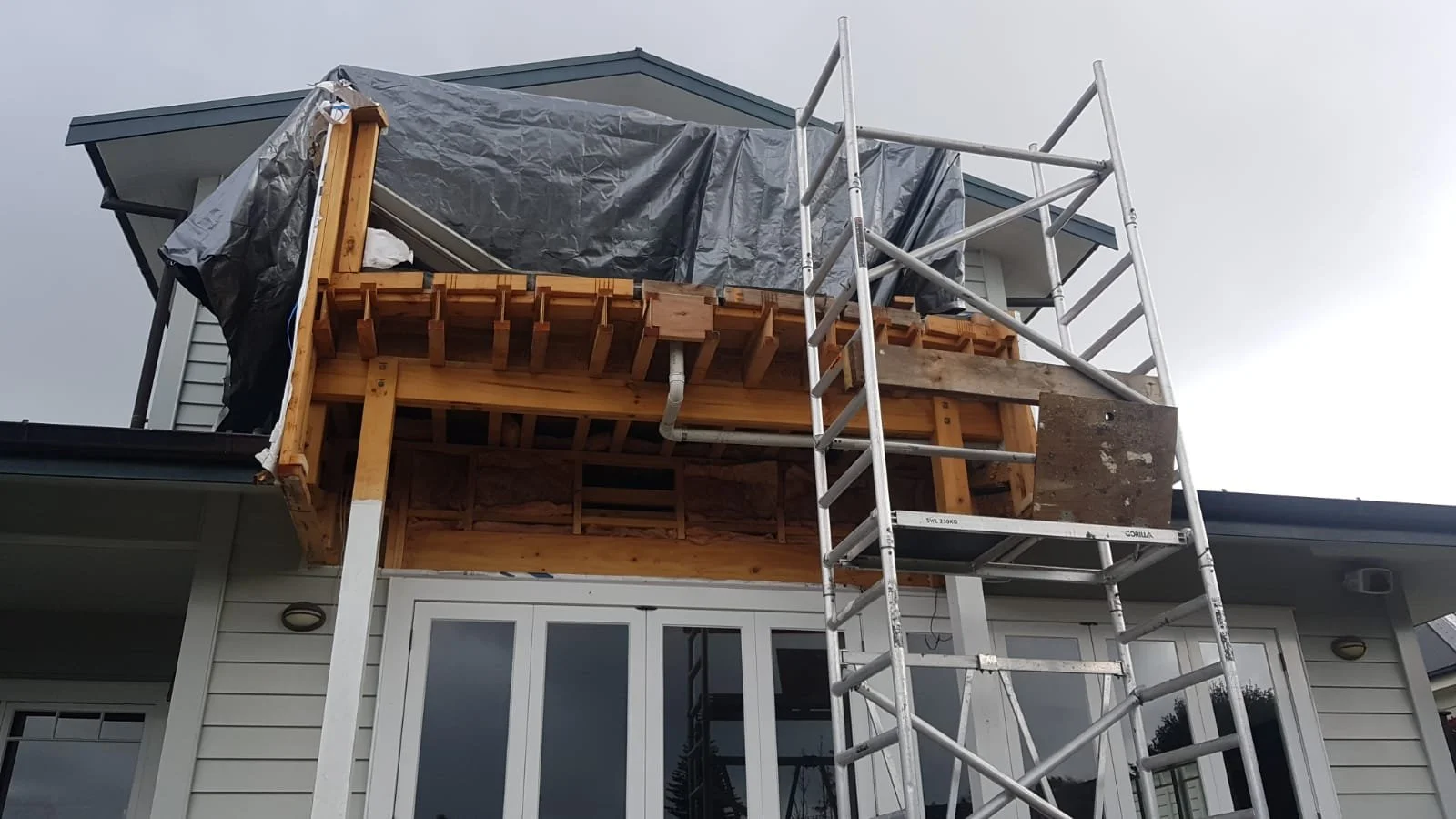 Construction scaffolding next to a house with part of the balcony under renovation, covered with a tarp, and showing exposed wooden framing.
