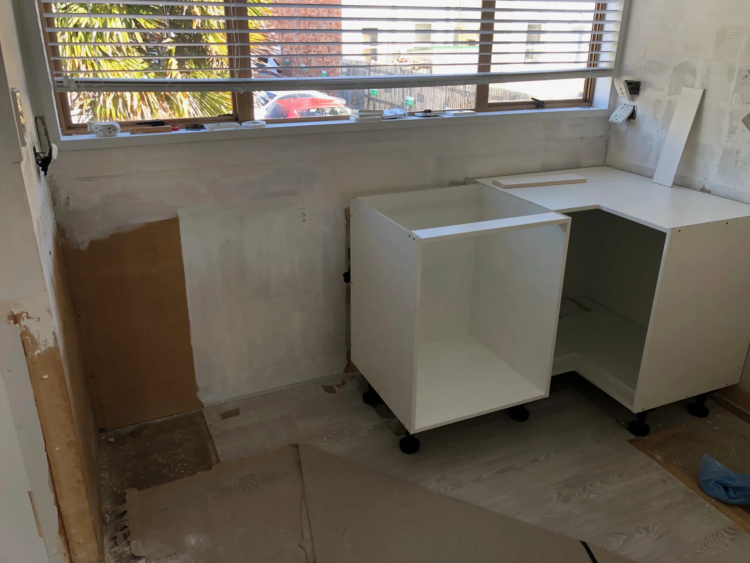 Interior of a room under renovation with partially assembled white cabinets, a window with blinds, and construction debris on the floor.