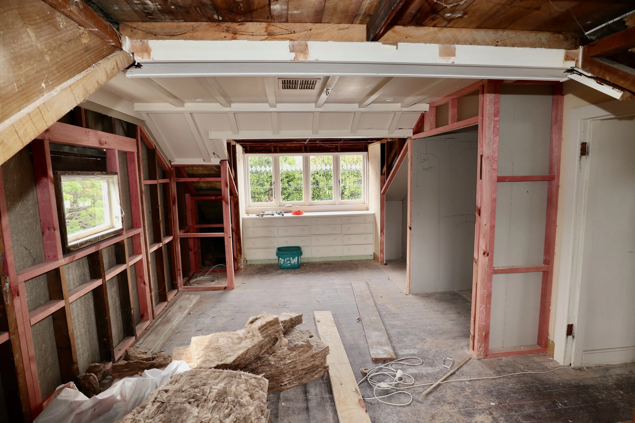 Room under renovation with exposed wooden framing, insulation on the floor, and a window at the far end. Construction materials and tools are scattered around.