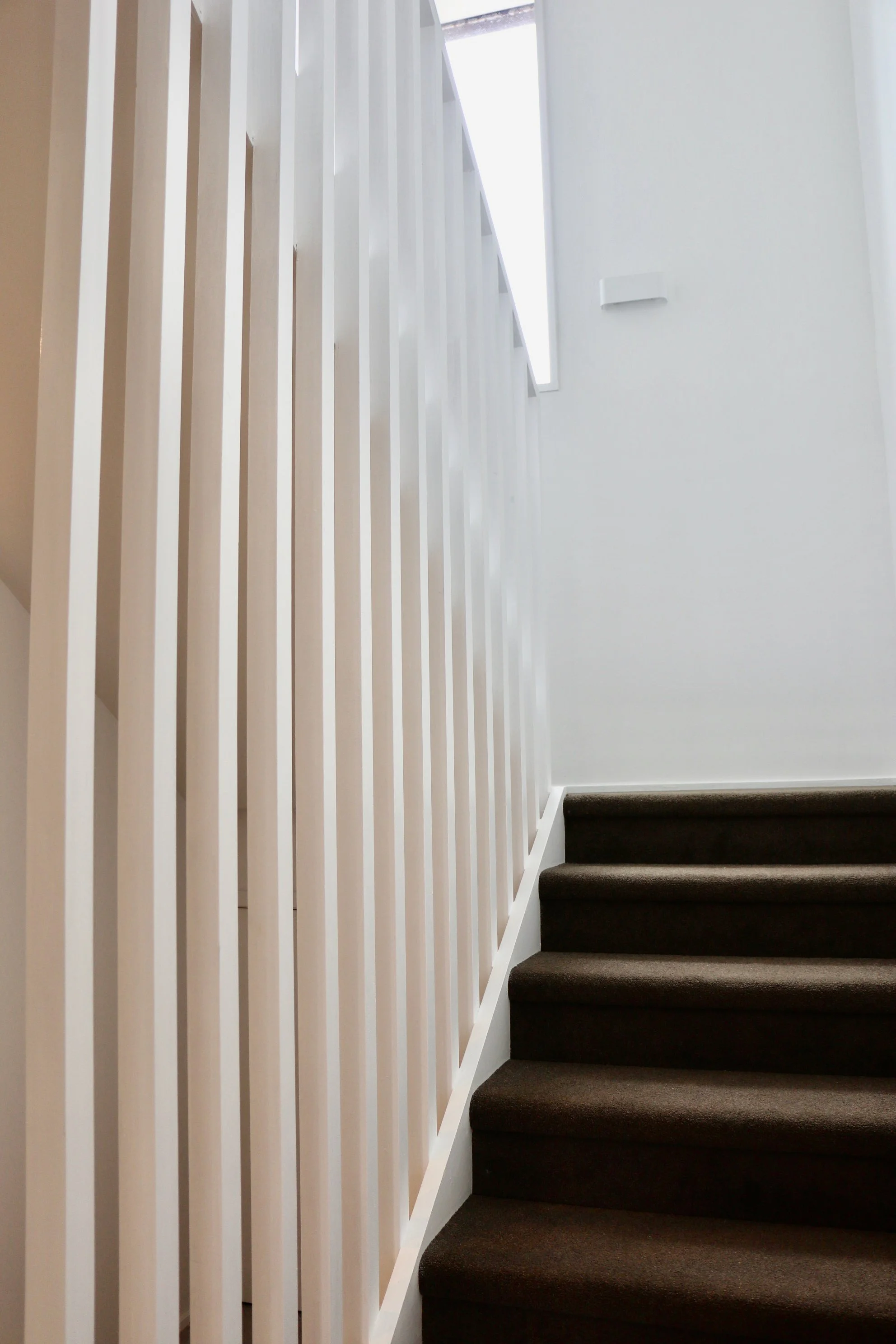 Interior view of a staircase with dark carpeted steps, white wall, and a white railing made of vertical slats. Natural light coming from a narrow window or opening above.