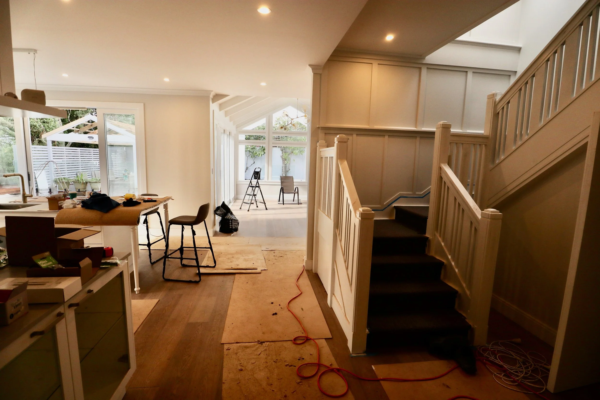Interior view of a house under renovation with scattered tools and furniture, showing a staircase on the right and a large window area at the back.