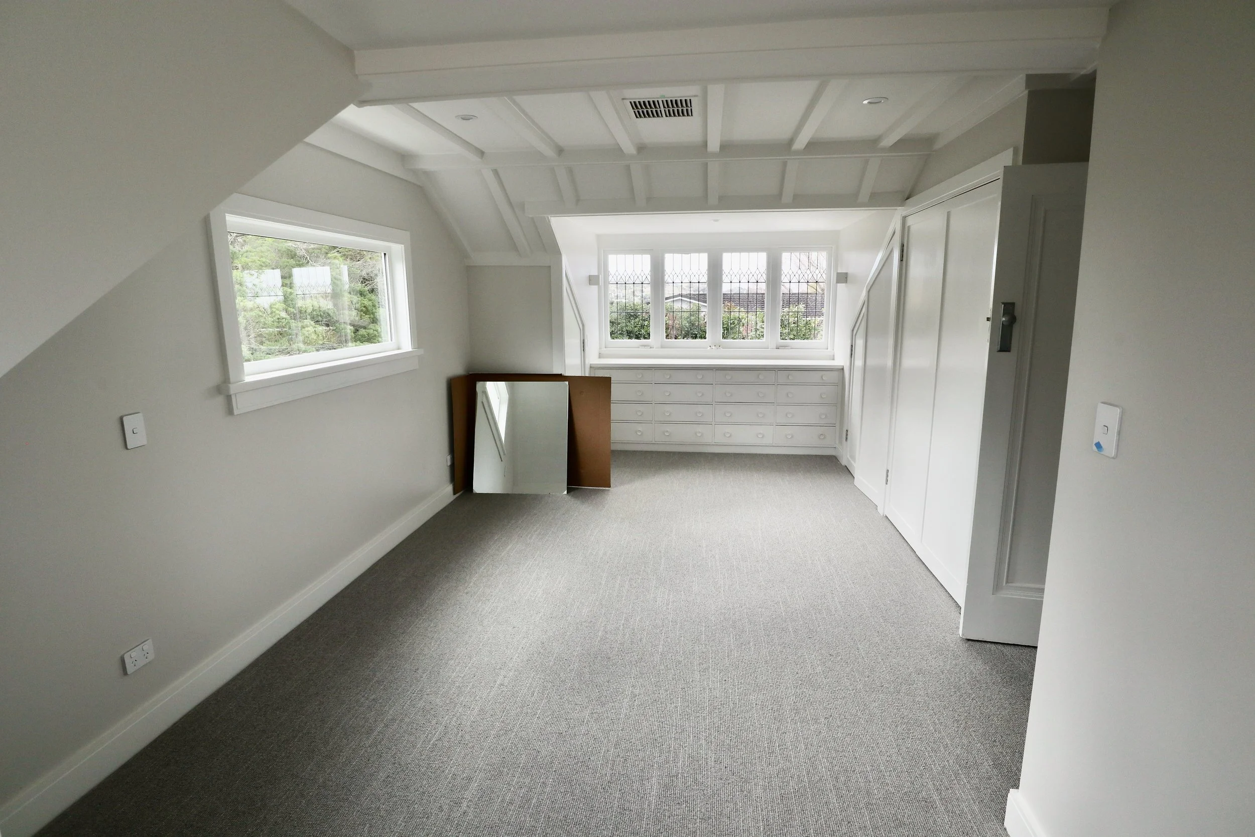 Empty attic room with white walls, gray carpet, built-in white drawers beneath a large window, and a small rectangular mirror leaning against the wall.