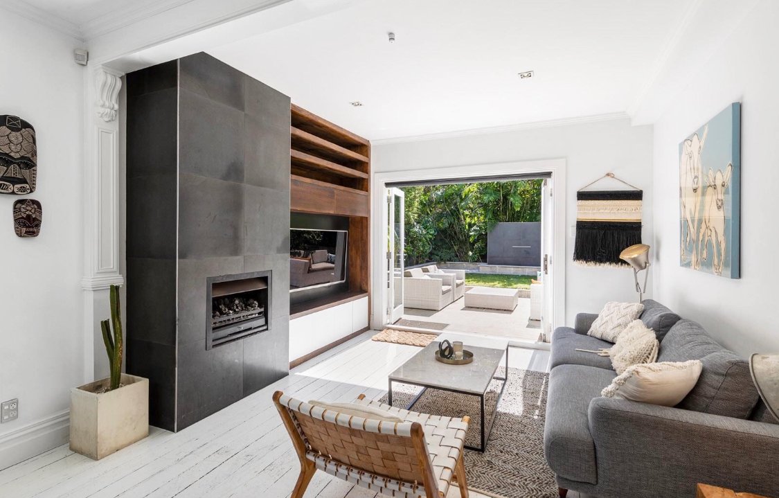 Living room with a gray sofa, a wooden and metal coffee table, a patterned rug, a wall-mounted fireplace, a built-in wooden shelf, and sliding glass doors opening to a patio with outdoor seating and greenery.