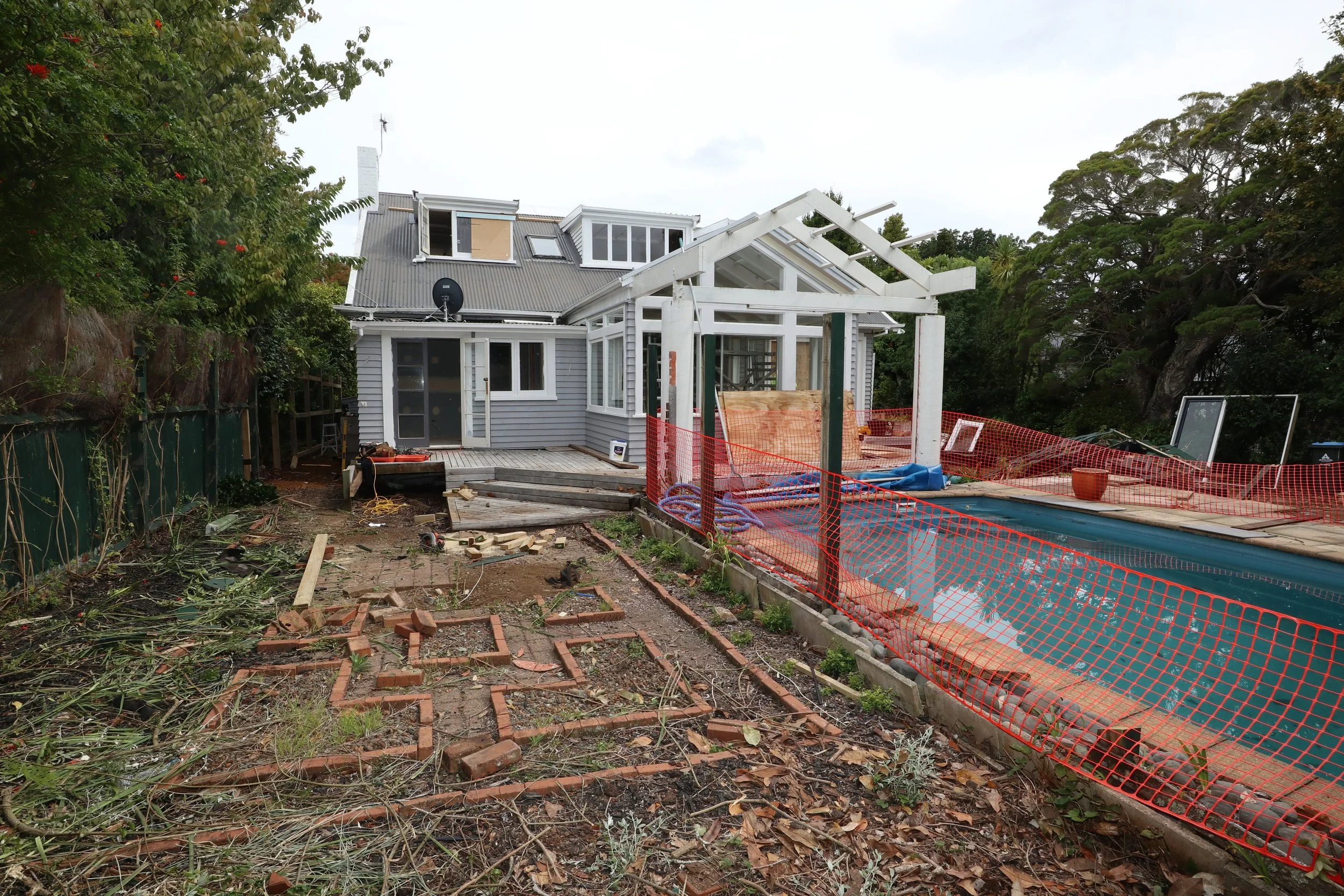 A house under renovation with a swimming pool covered with a tarp, construction materials and orange safety fencing around the pool, and a partially built structure near the house.