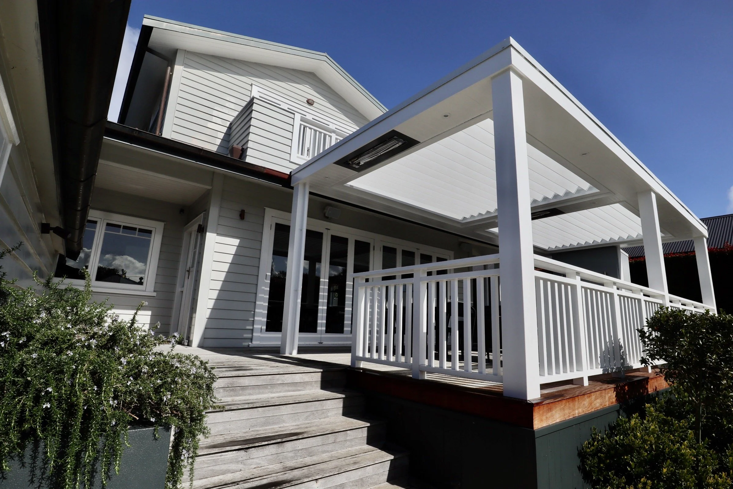 Exterior view of a white house with a wooden deck and stairs, fenced with white railing, under a blue sky.