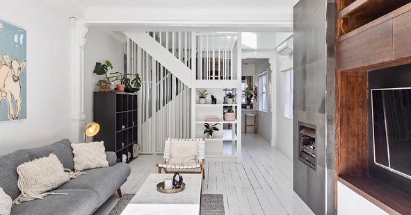 Living room with gray sofa, white armchair, black bookshelf, plants, and a staircase with white railing.