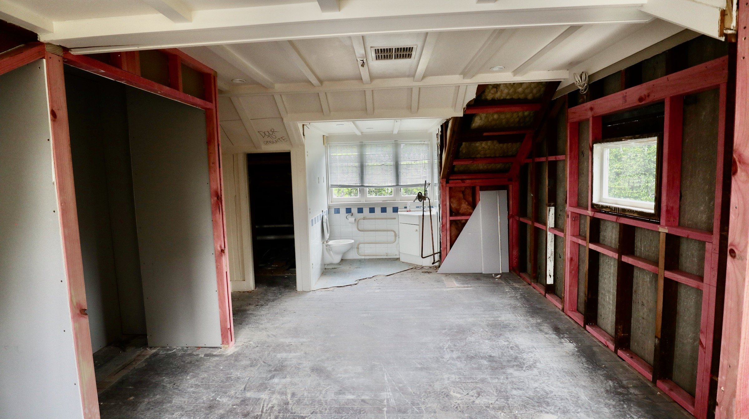 Room under renovation with exposed wooden studs on walls, a window with blinds, a damaged floor, and a bathroom with a toilet in the background.