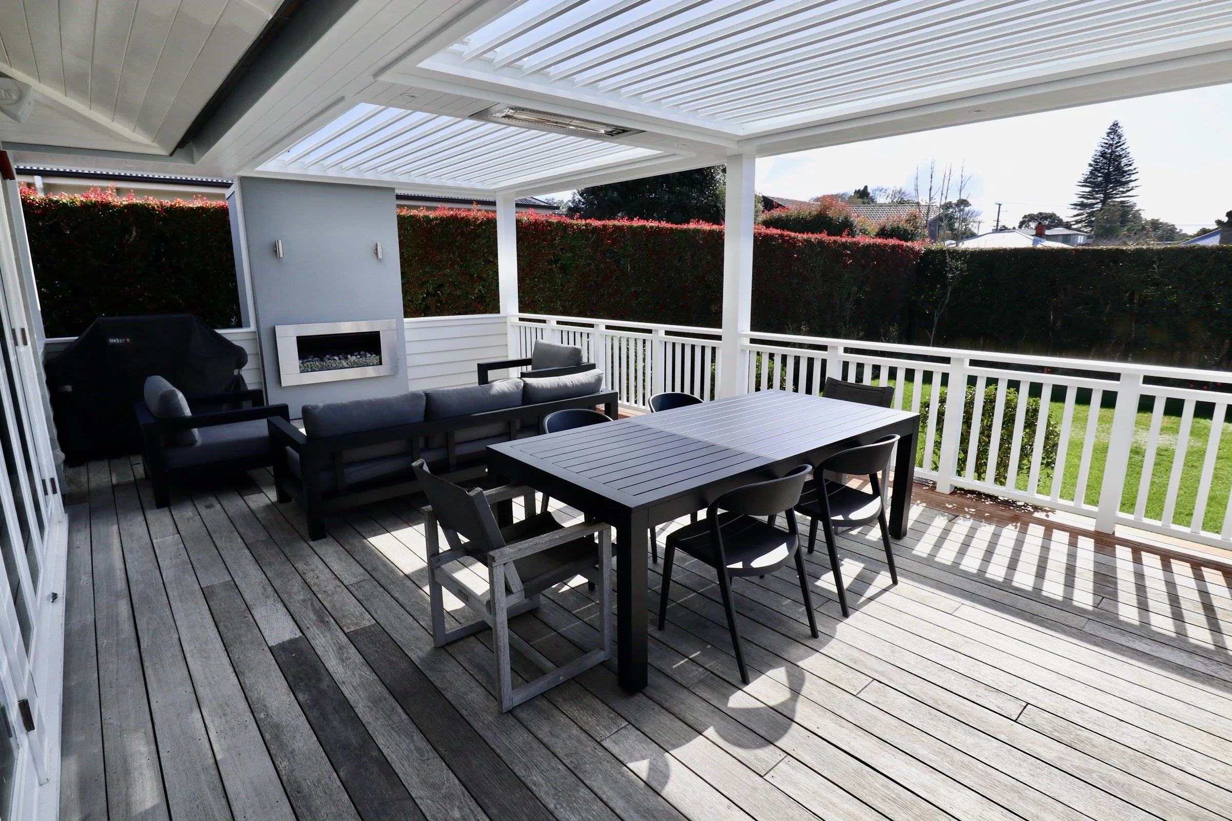 Covered outdoor patio with wooden deck, black dining table with six chairs, seating area with black and gray furniture, and a fireplace, overlooking a backyard with a green lawn and tall hedge.