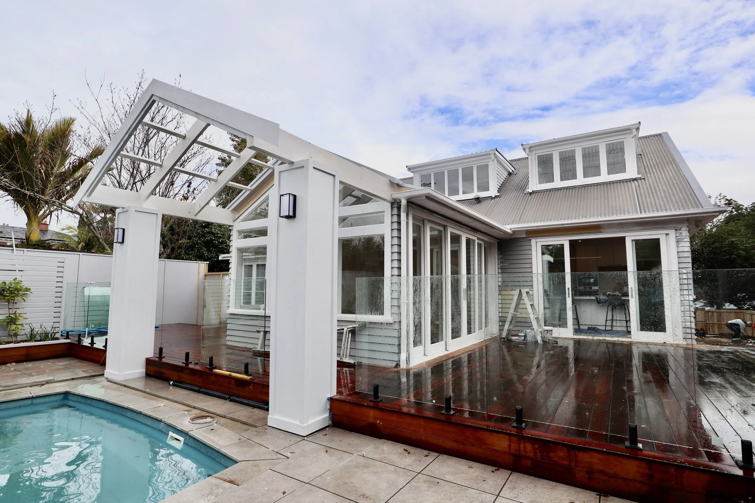Backyard of a house with a pool, newly built glass fence, and a wooden deck on a rainy day.