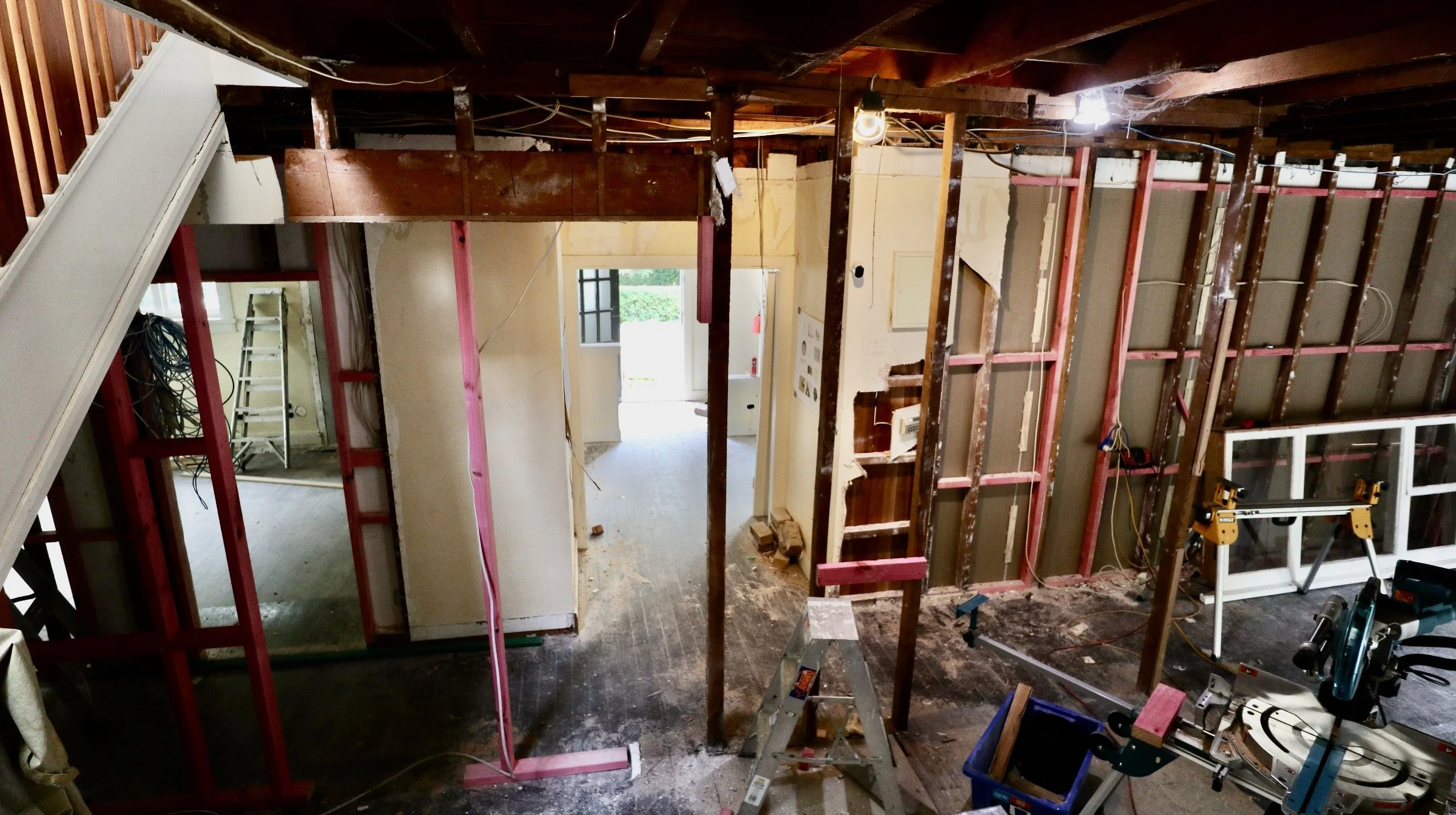 Interior view of a room under renovation with exposed wooden framing, construction tools, and equipment, including a ladder, saw, and work tables.
