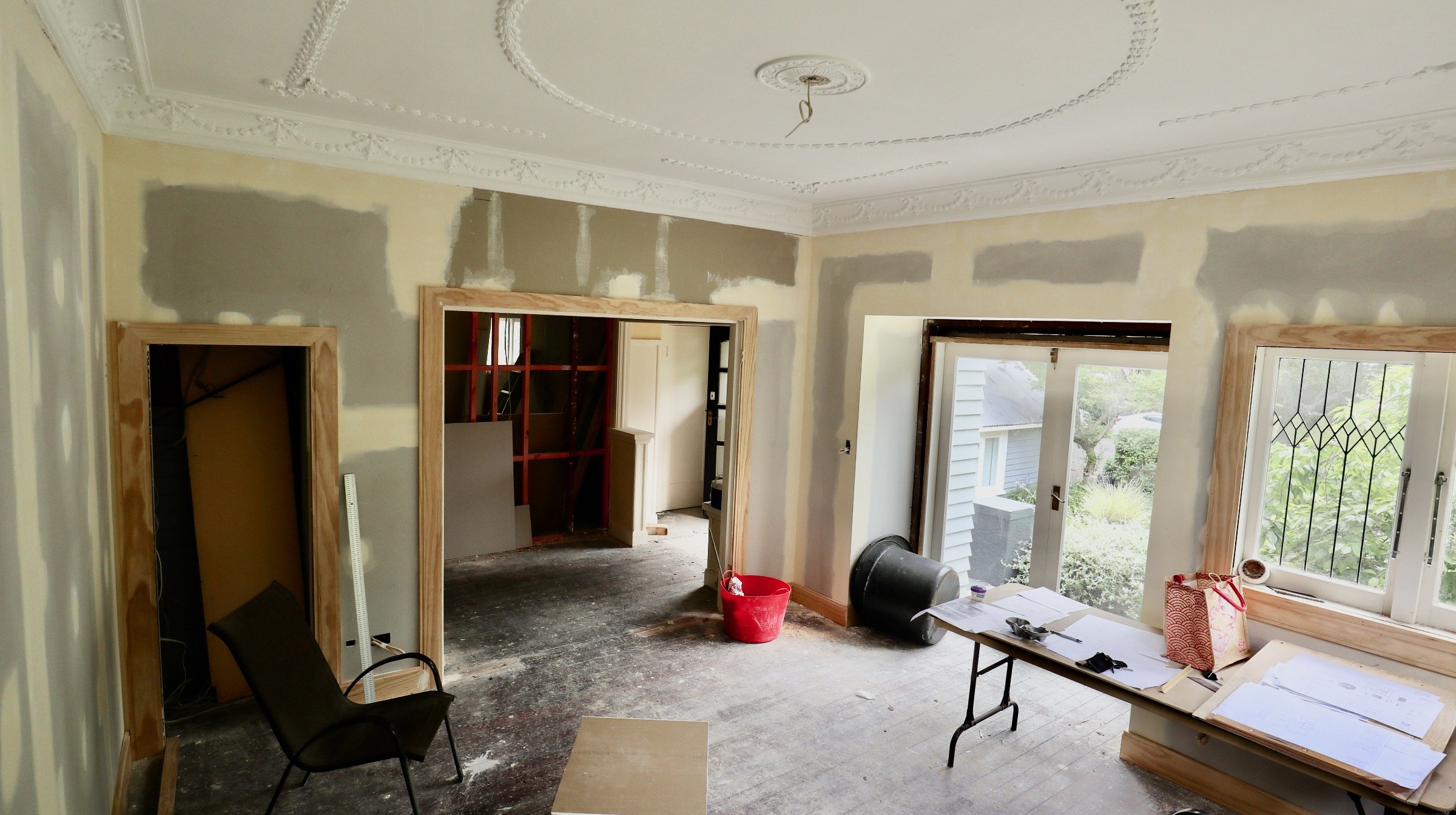 Interior of a house under renovation with unfinished walls, construction materials, a black chair, and windows with landscaped yard outside.