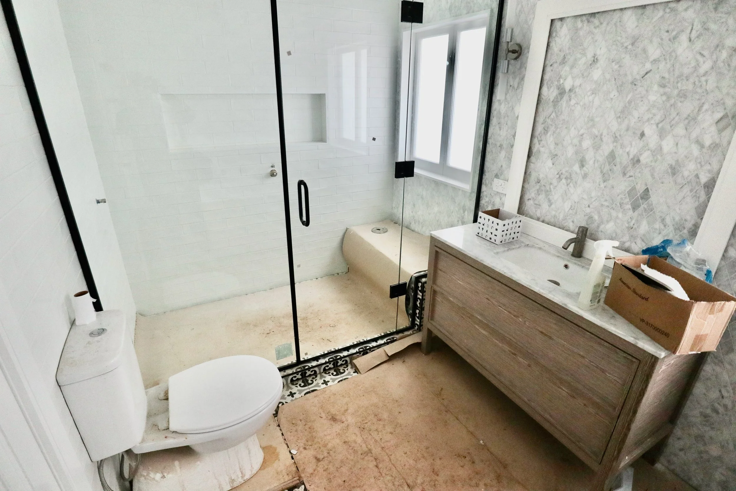 A bathroom under renovation featuring a glass shower enclosure, a toilet, a wooden vanity with a marble countertop and a sink, a cardboard box, and various construction items, with a window allowing natural light.