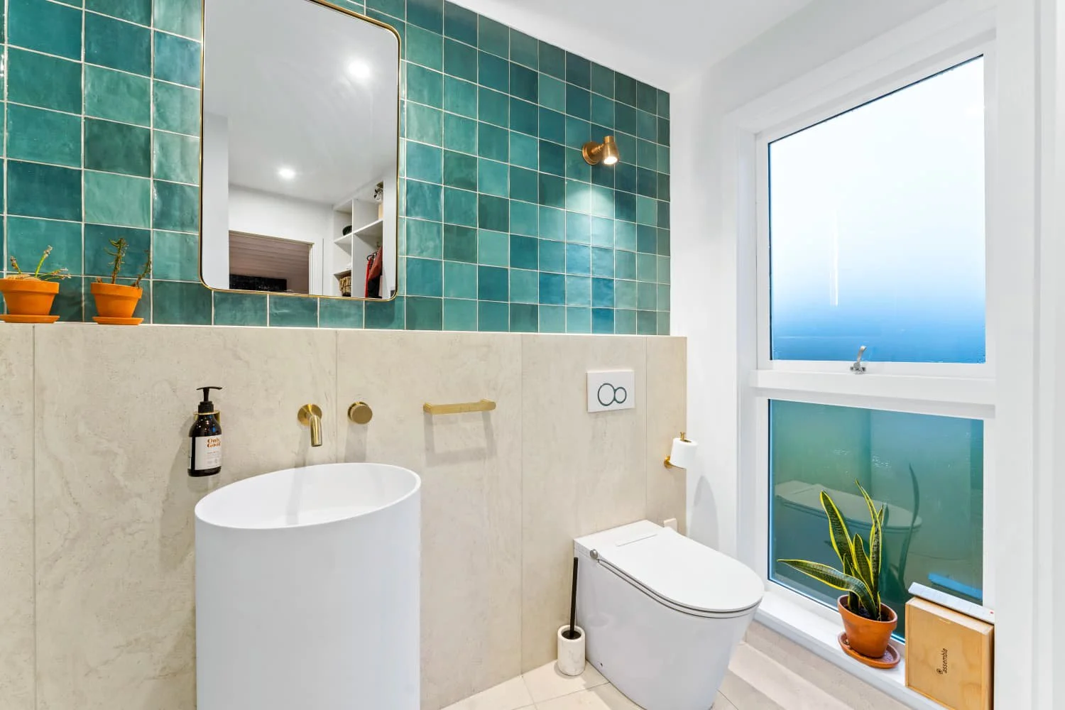 Modern bathroom with green tiled wall, white toilet, round white sink, a large mirror, a window with frosted glass, and potted plants.