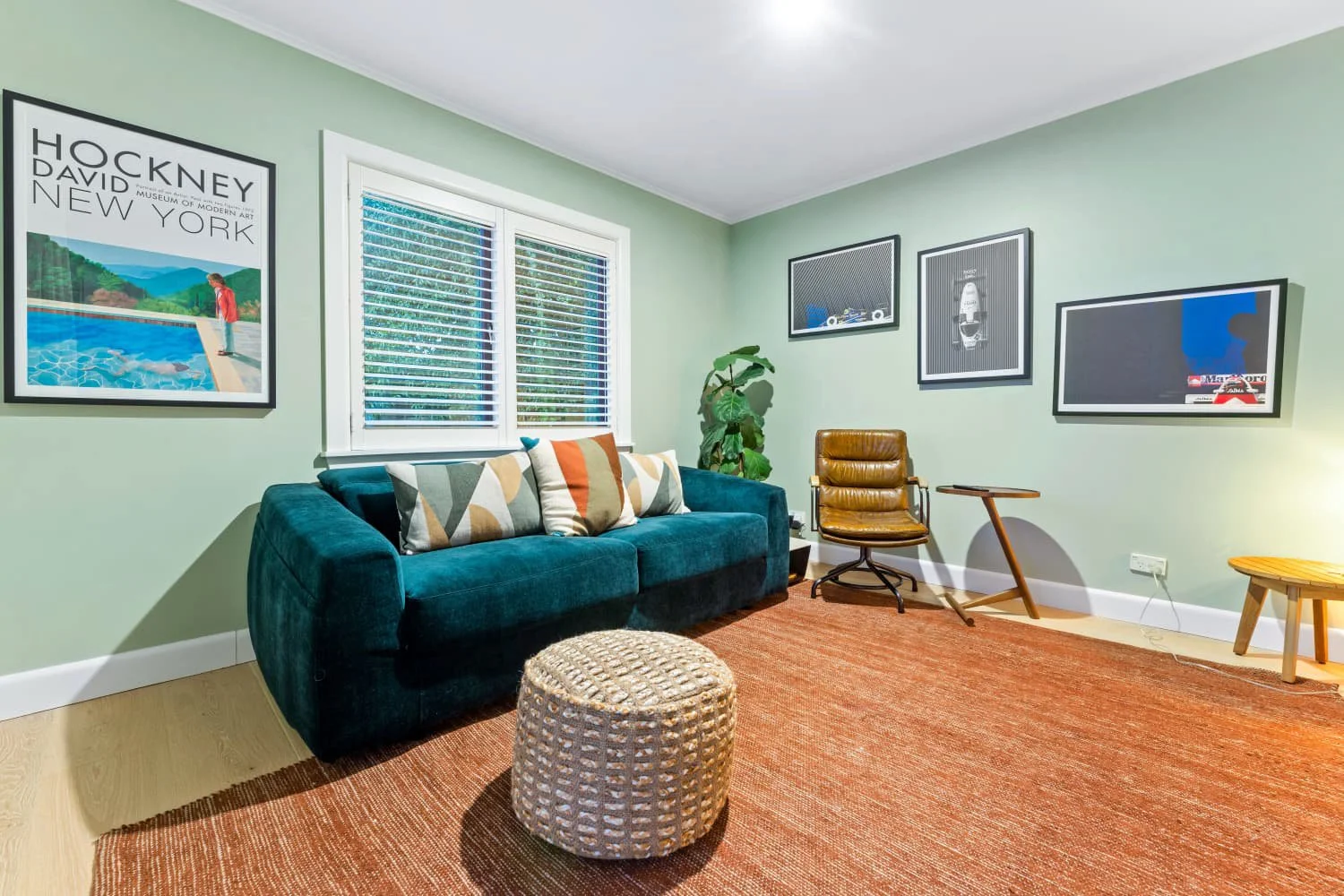 Living room with green walls, a teal sofa with patterned pillows, a round woven ottoman, a brown leather chair, a small wooden side table, framed artwork on the walls, a large window with blinds, and a potted plant.