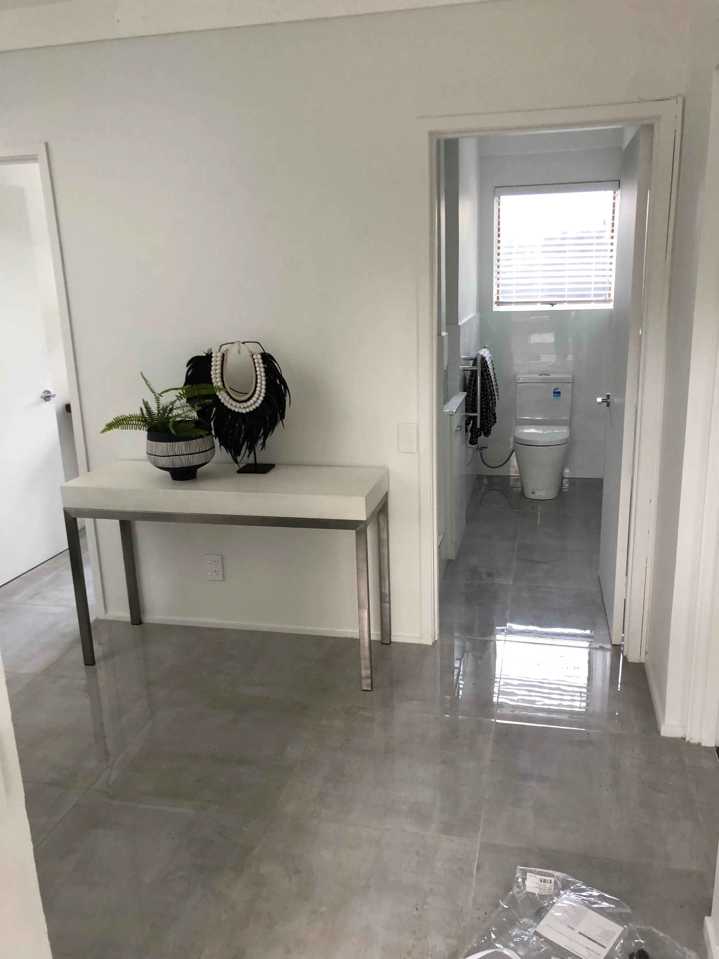 Interior of a modern home, showing a hallway with a small table decorated with a plant and jewelry display, leading to a bathroom with a toilet and window with blinds.
