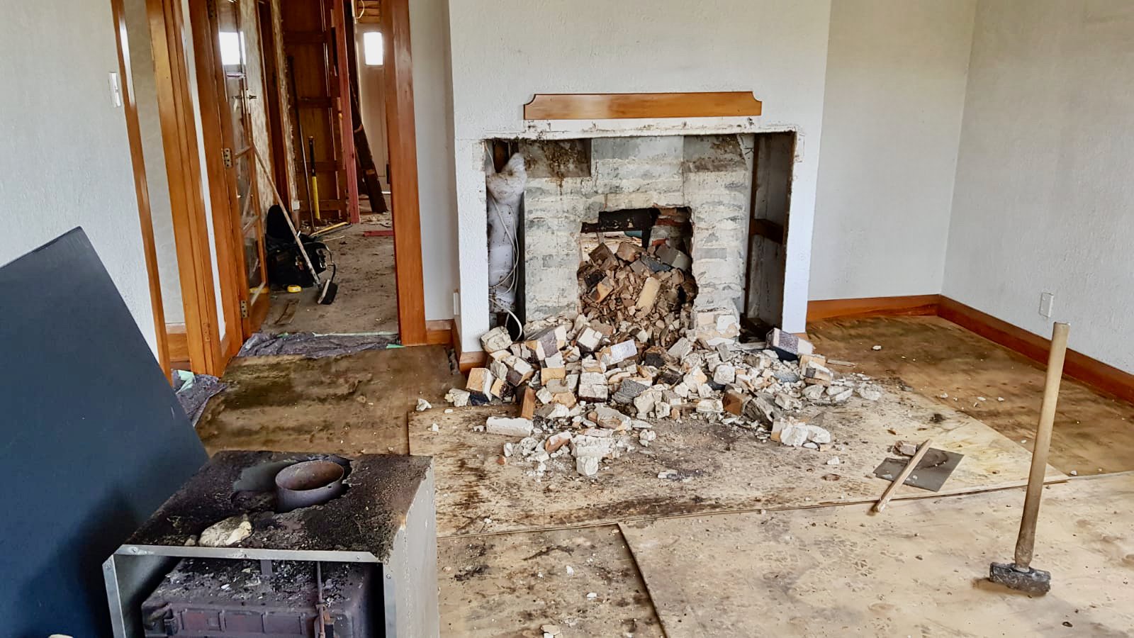 Living room under renovation with a partially demolished brick fireplace, scattered debris, construction tools, and exposed floor.