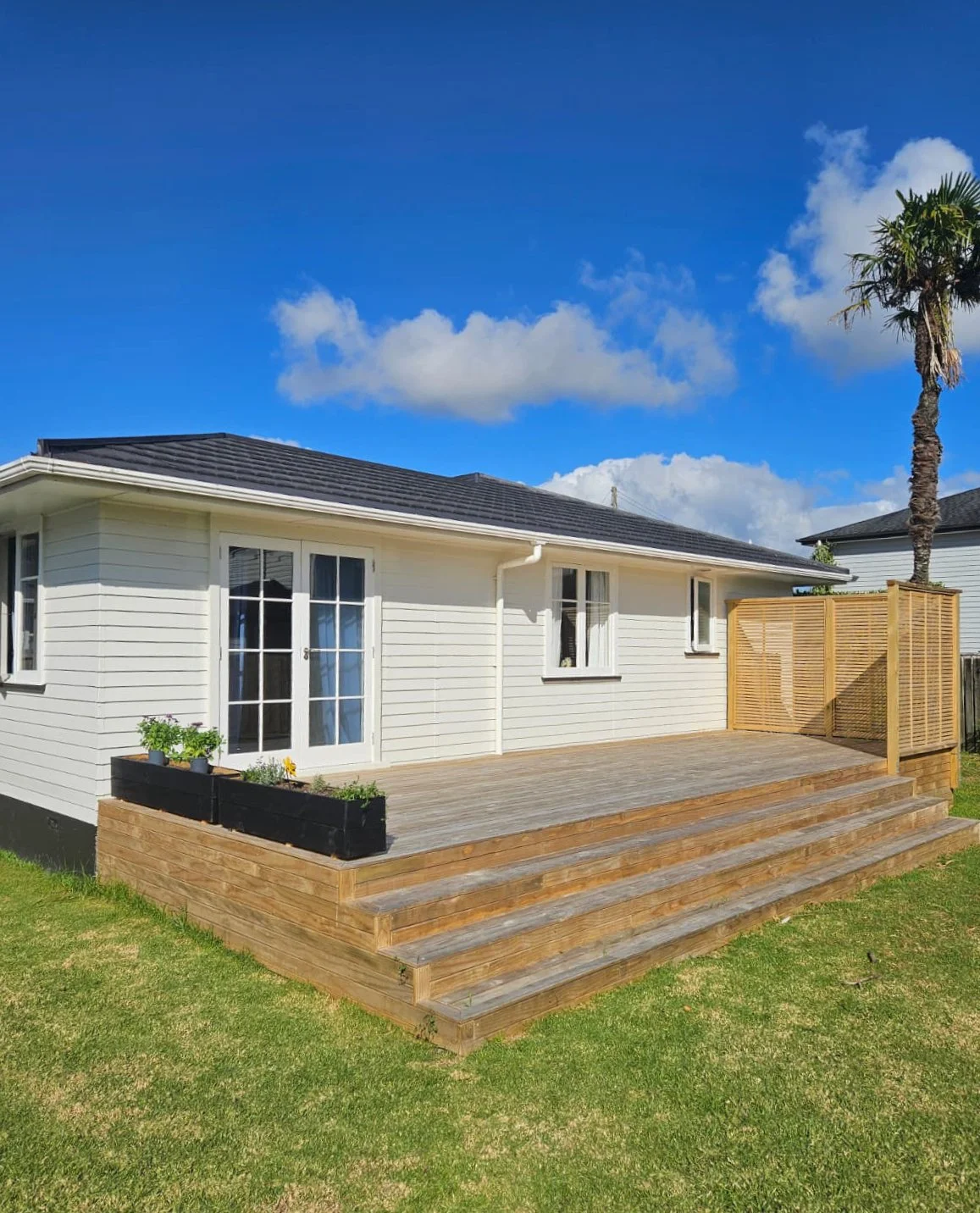 Backyard with wooden deck, white house, sliding glass doors, small flower bed, grass lawn, palm tree, blue sky with clouds.