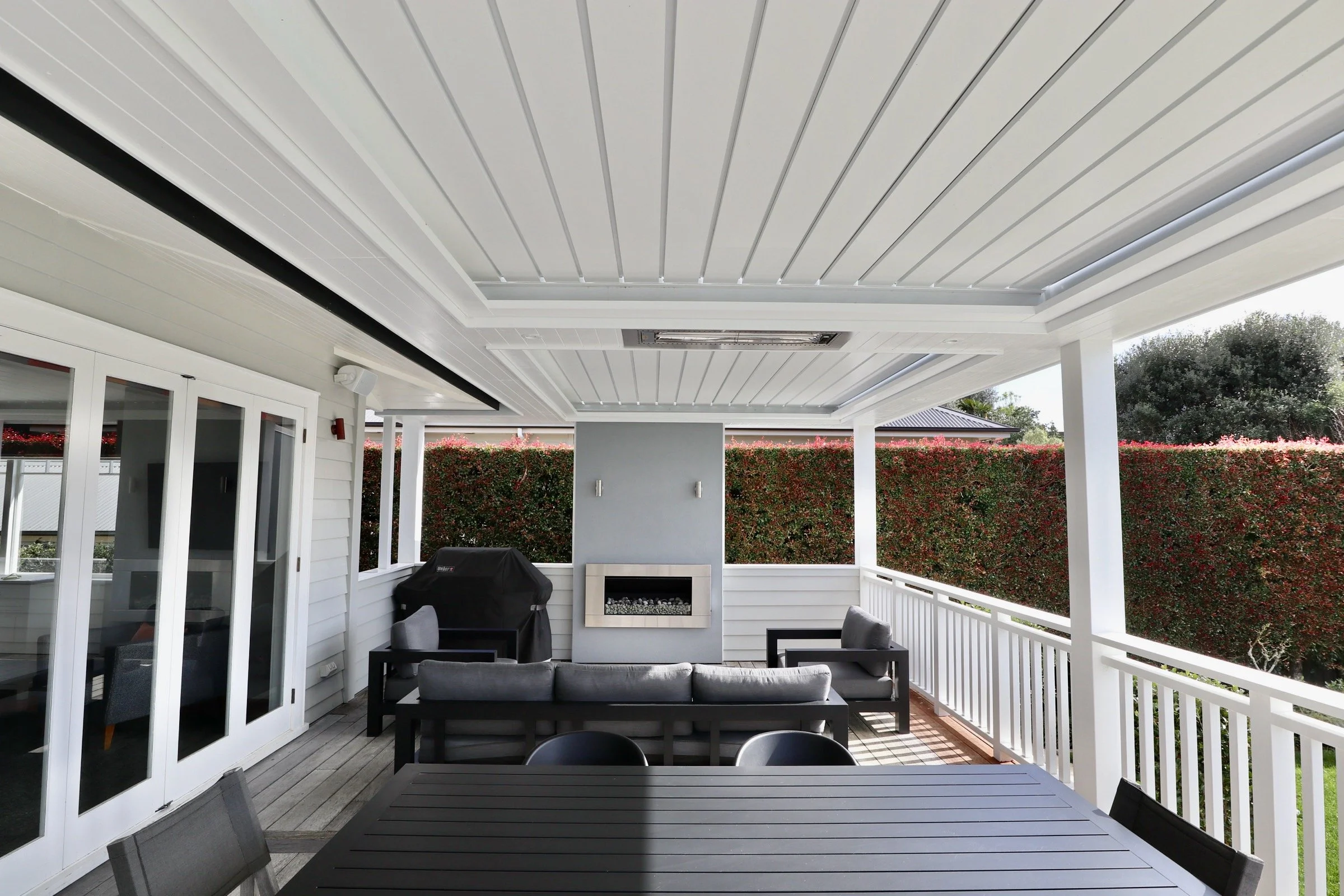Covered backyard patio with white ceiling and railing, black outdoor furniture, grill covered with black cover, and fireplace on a blue wall, overlooking a tall hedge.