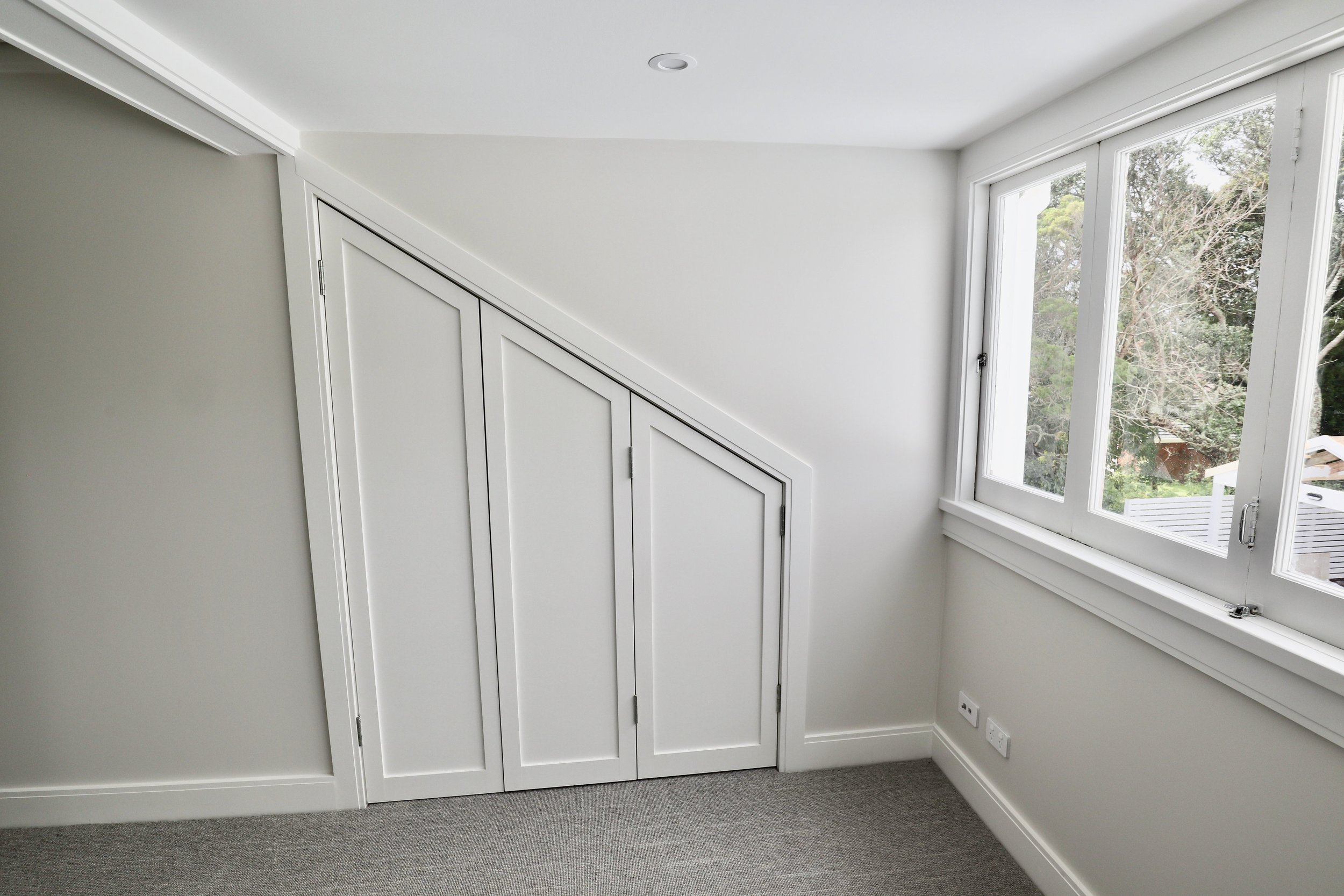 Empty room with a sloped ceiling, white walls, small built-in storage cabinets, and large windows showing a view of trees outside.