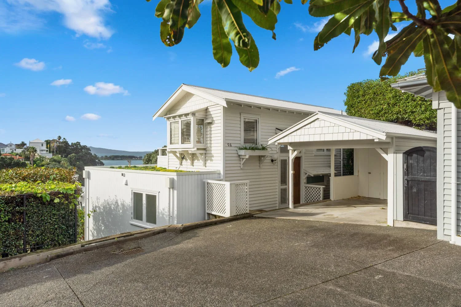A white two-story house with a gabled roof and a small balcony, set against a background of blue sky, greenery, and distant water. The house has shuttered windows, a black door, and a white fence, with leafy trees framing the scene.