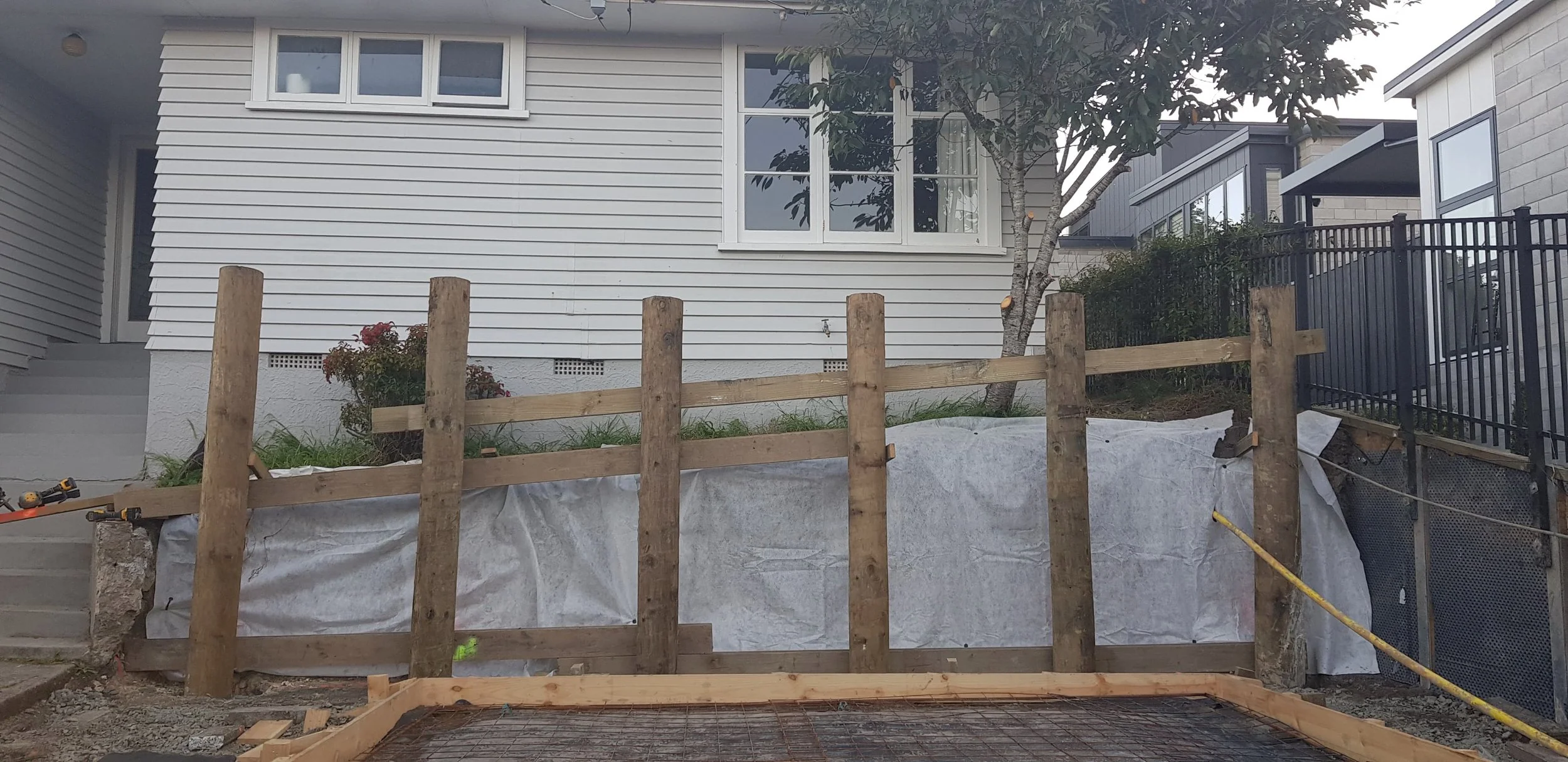 Construction site in front of a house with wooden fence posts, partially built wooden fence, and construction materials. The house has white siding, windows, and steps leading to the front door. There is a small tree and some bushes nearby.