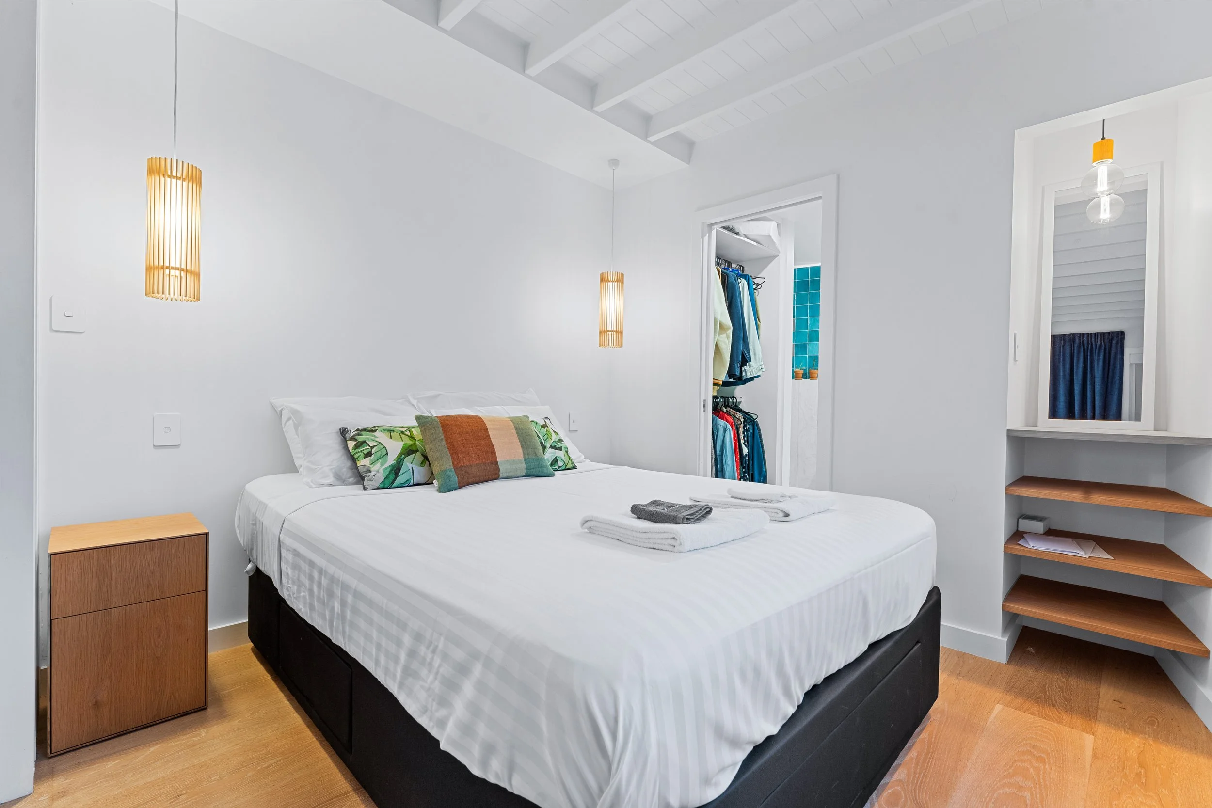 A clean, minimalist bedroom with a neatly made bed, white bedding, and decorative pillows. Wooden nightstand to the left, open closet with hanging clothes, and a mirror with a small shelf below on the right. Hanging pendant lights with warm glow on e