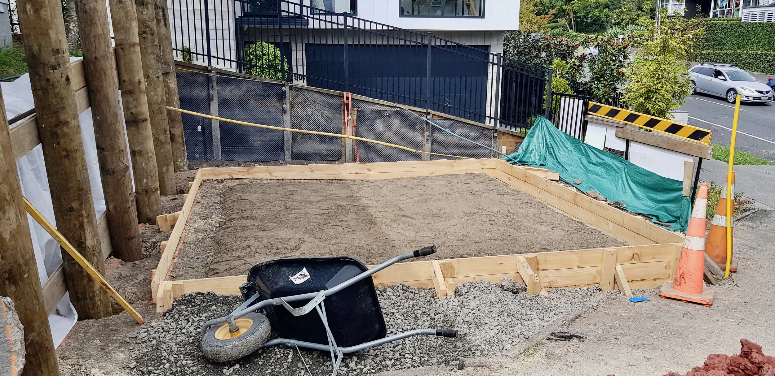 Construction site next to sidewalk with wooden frame, wheelbarrow, and orange safety cones.