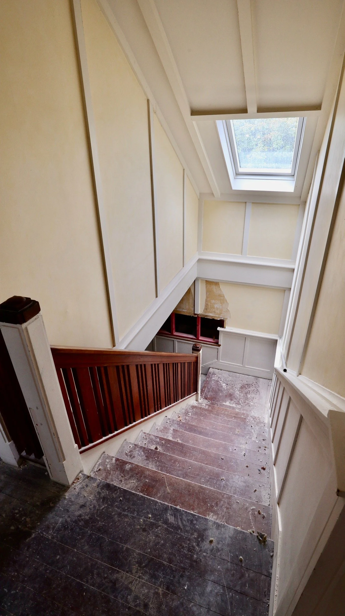 View down a staircase showing worn wooden steps, a partial newel post, and a large skylight window overhead, with light yellow wall panels and a white ceiling.