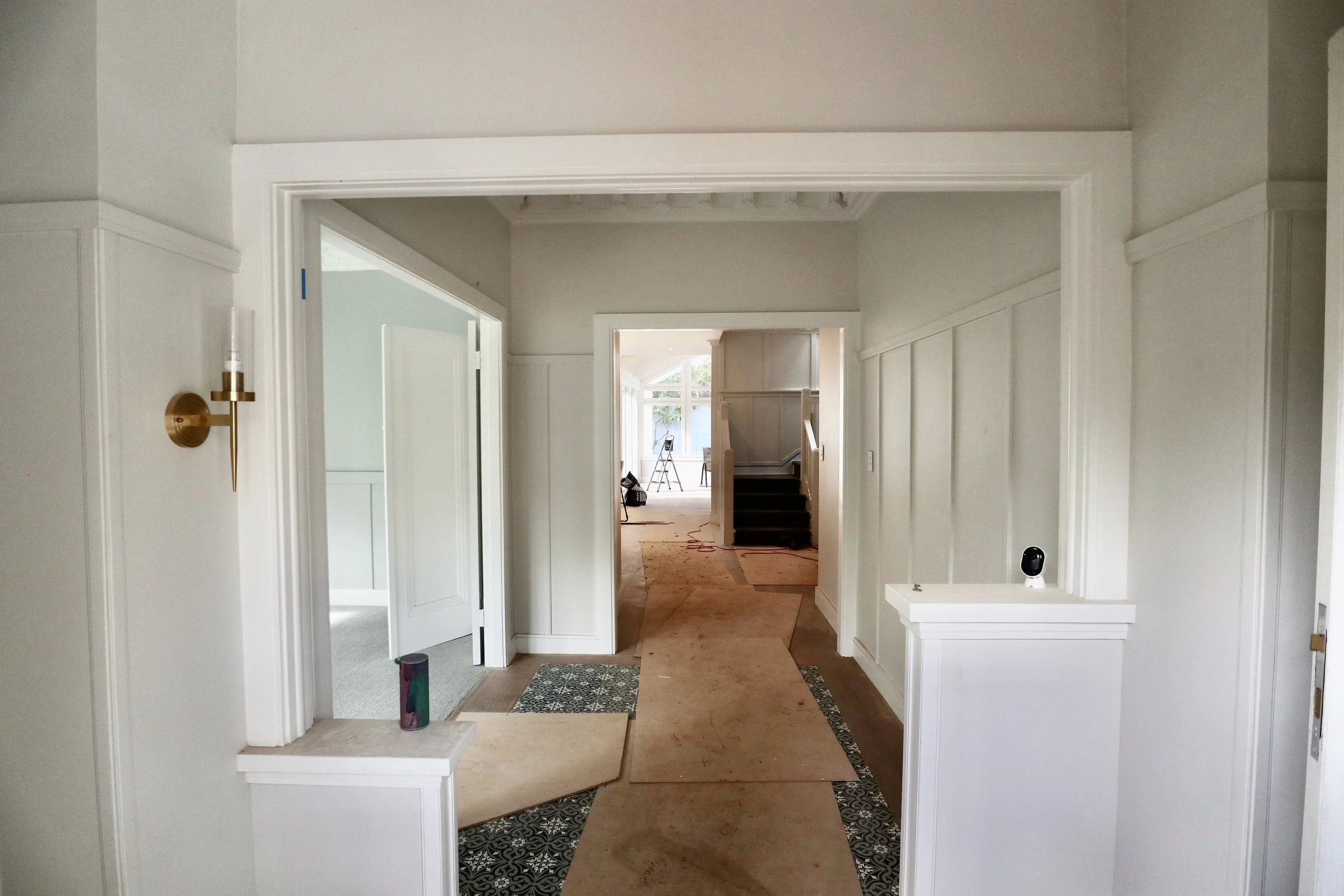 Interior view of a house under renovation, showing a hallway with Masonite boards on the floor and tools in the background.