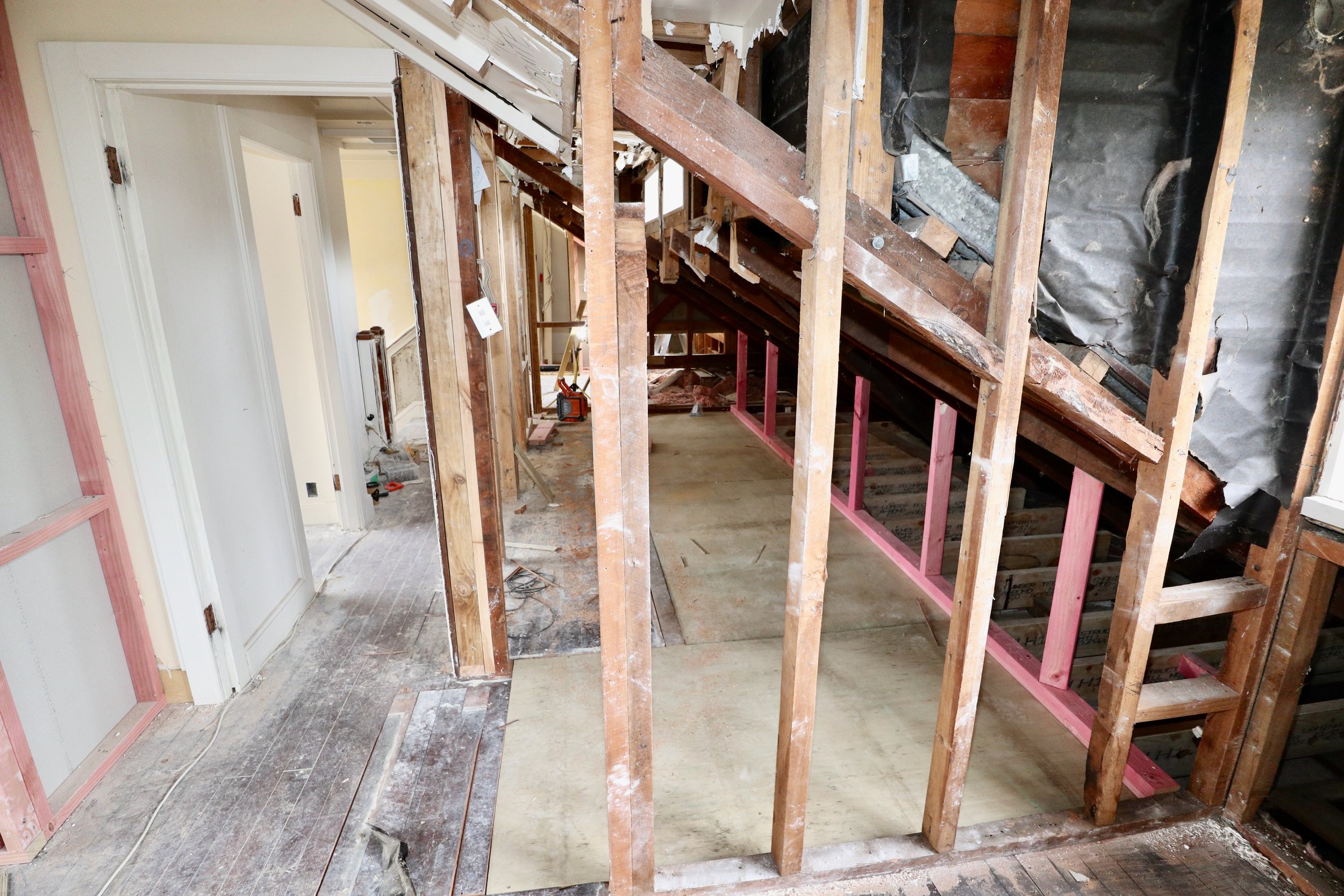 Interior of a house under renovation, showing exposed wooden framing, partially removed drywall, and construction tools on the floor.