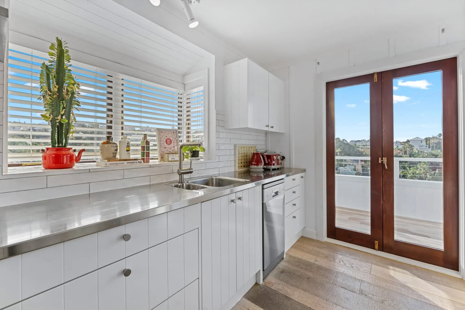 A bright, modern kitchen with white cabinets, a stainless steel countertop, a small sink, and a window with white blinds showing a sunny outdoor view. There are red small appliances and kitchen items on the counter, and wooden French doors lead to an