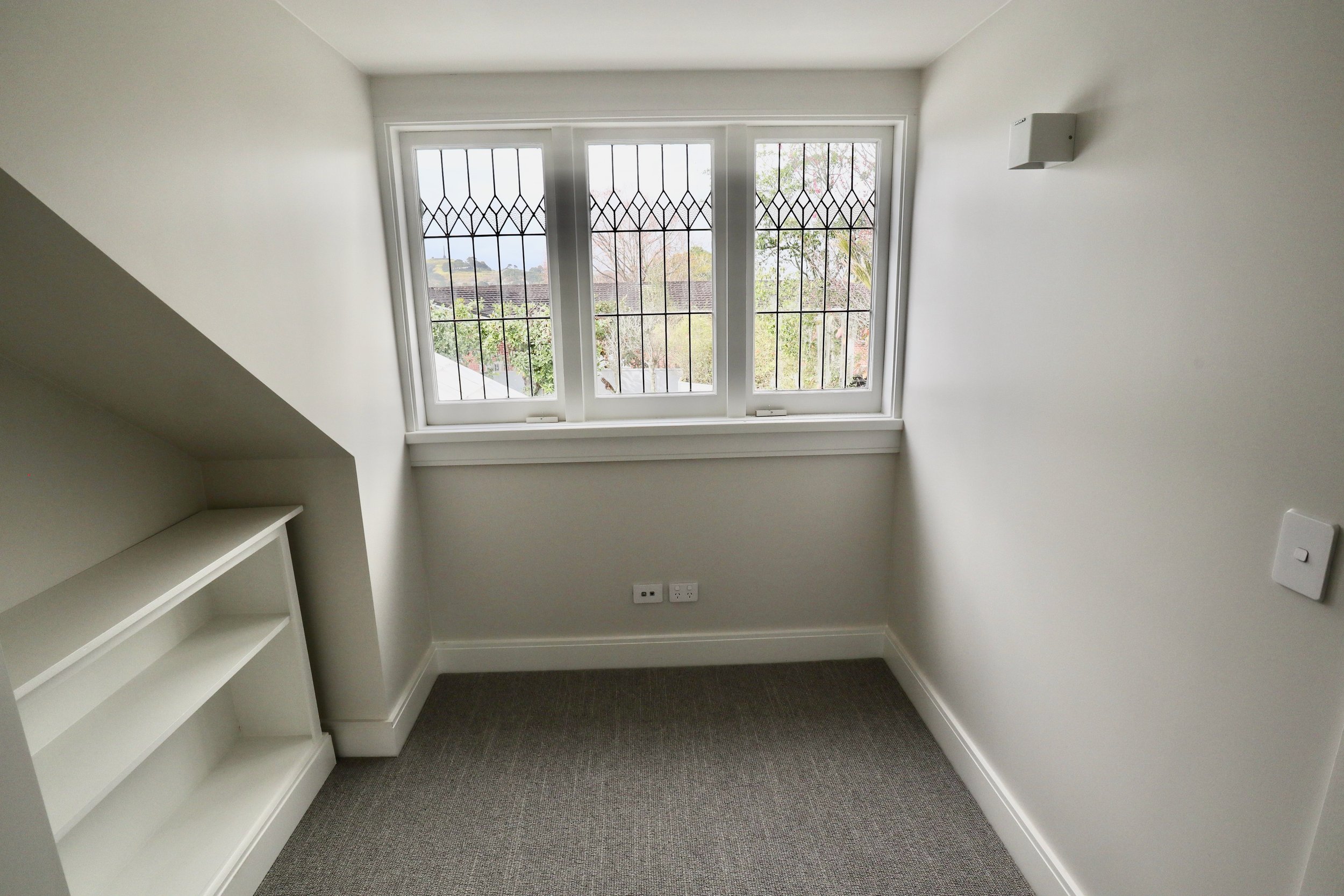 Empty small room with a large window, gray carpet, white walls, built-in white shelving, and electrical outlets.