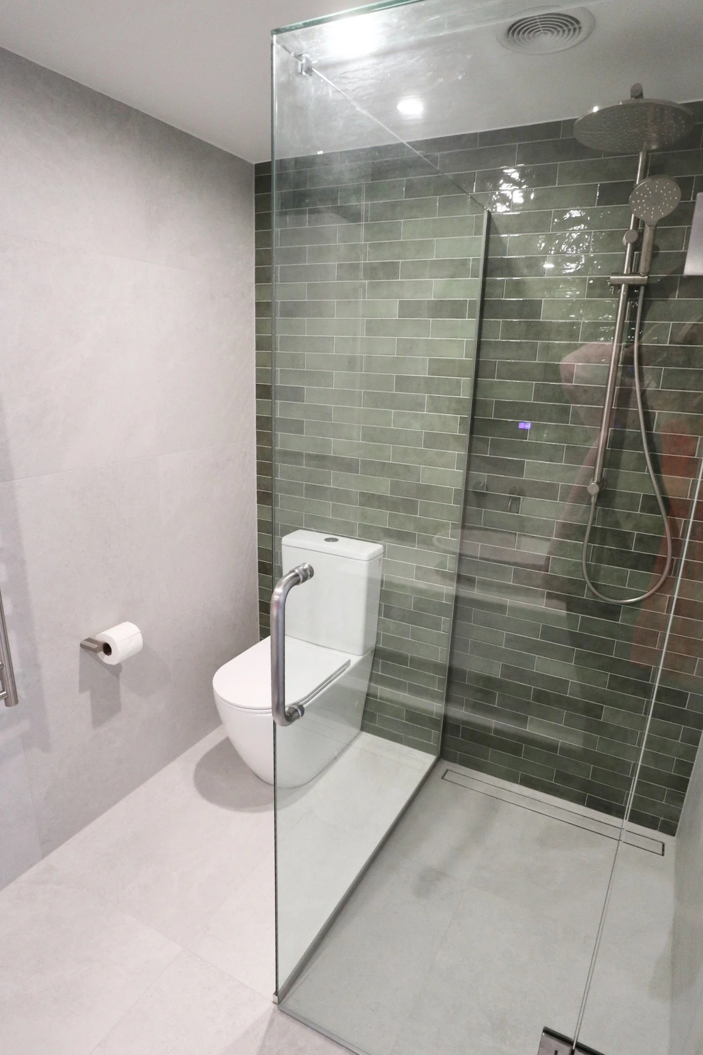 Modern bathroom with a transparent glass shower enclosure, gray brick tile wall, white toilet, and toilet paper holder.