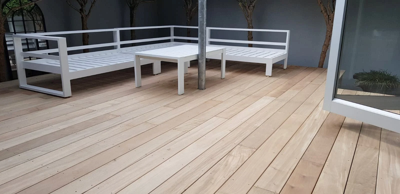 An outdoor wooden deck with white metal patio furniture including a long corner sofa, a small table, and a larger table, with a sliding glass door to the right and trees visible in the background.