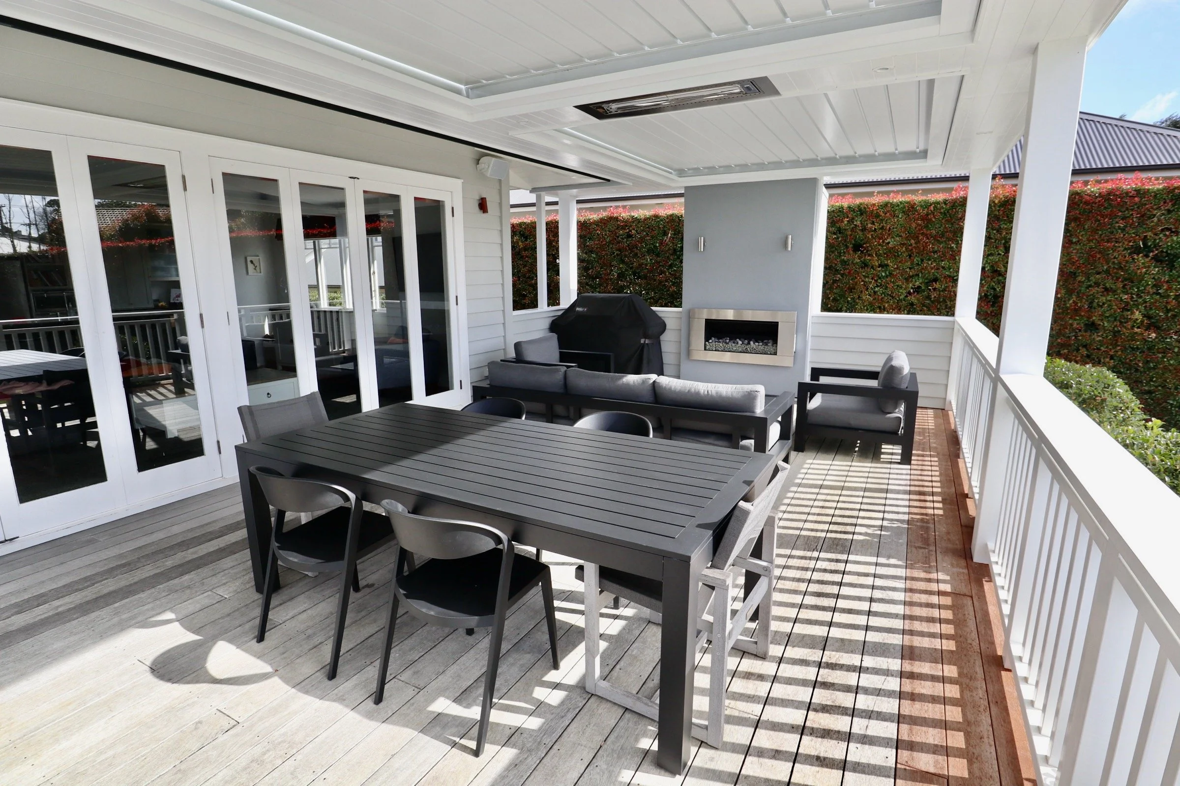 Outdoor covered patio with black dining table and chairs, a gray sofa with cushions, a cushioned armchair, a gas fireplace built into a gray wall, a grill covered with a black cover, and white railing and siding with sliding glass doors leading insid