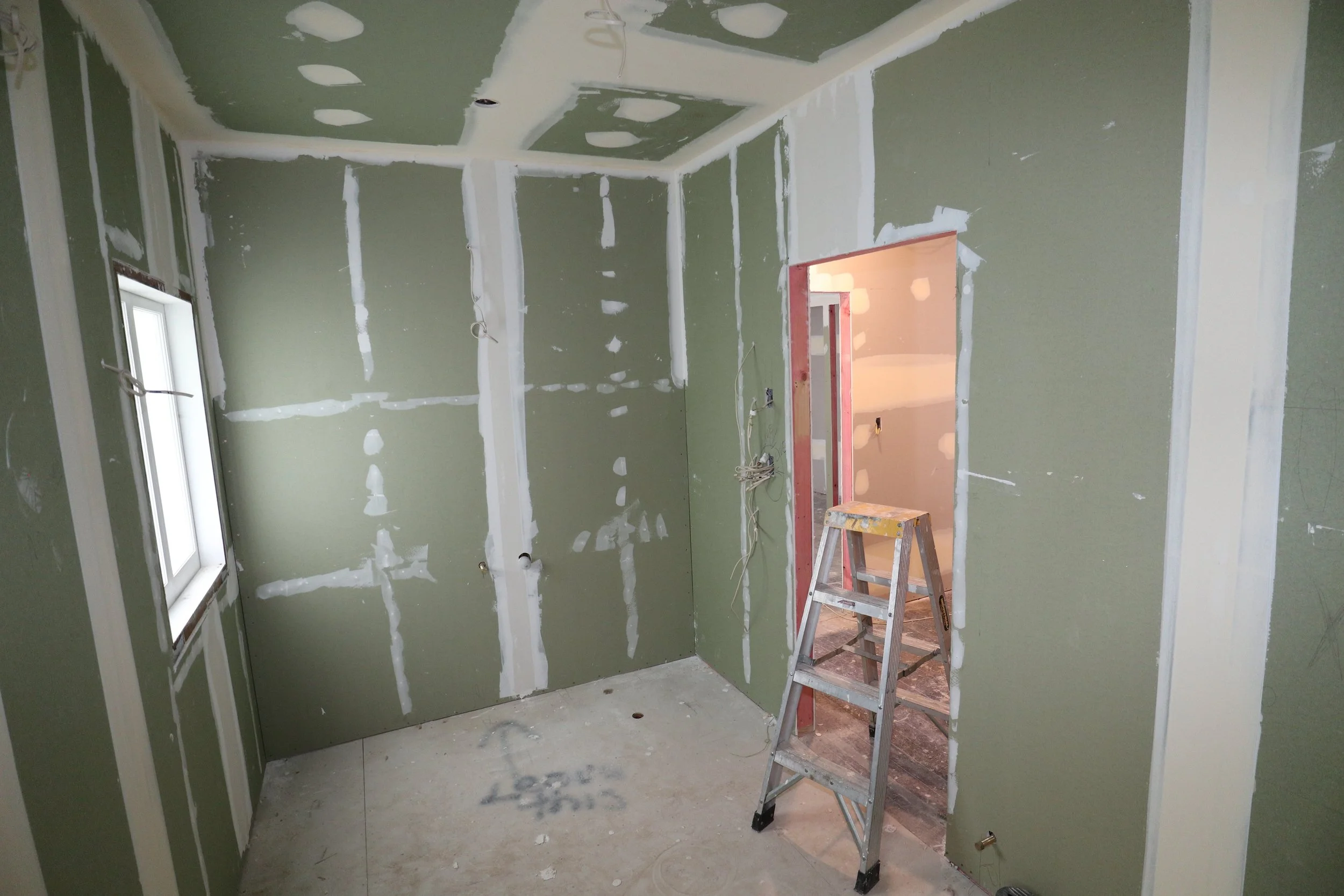 Interior of a room under construction with green drywall, a window on the left, and an open doorway on the right. There is a ladder standing in the middle of the room, and the walls and ceiling are partially finished with visible joint compound and d