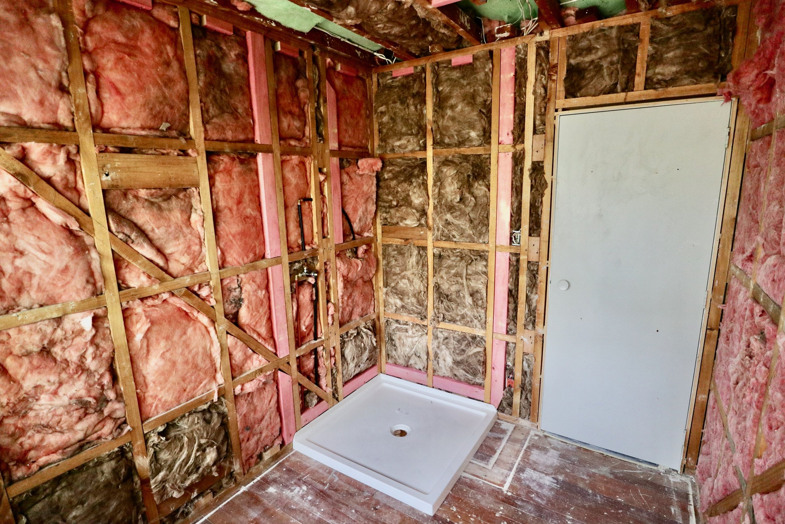 A bathroom under renovation with exposed wall studs, pink insulation, and a white shower base installed. A white door is closed on the right side.
