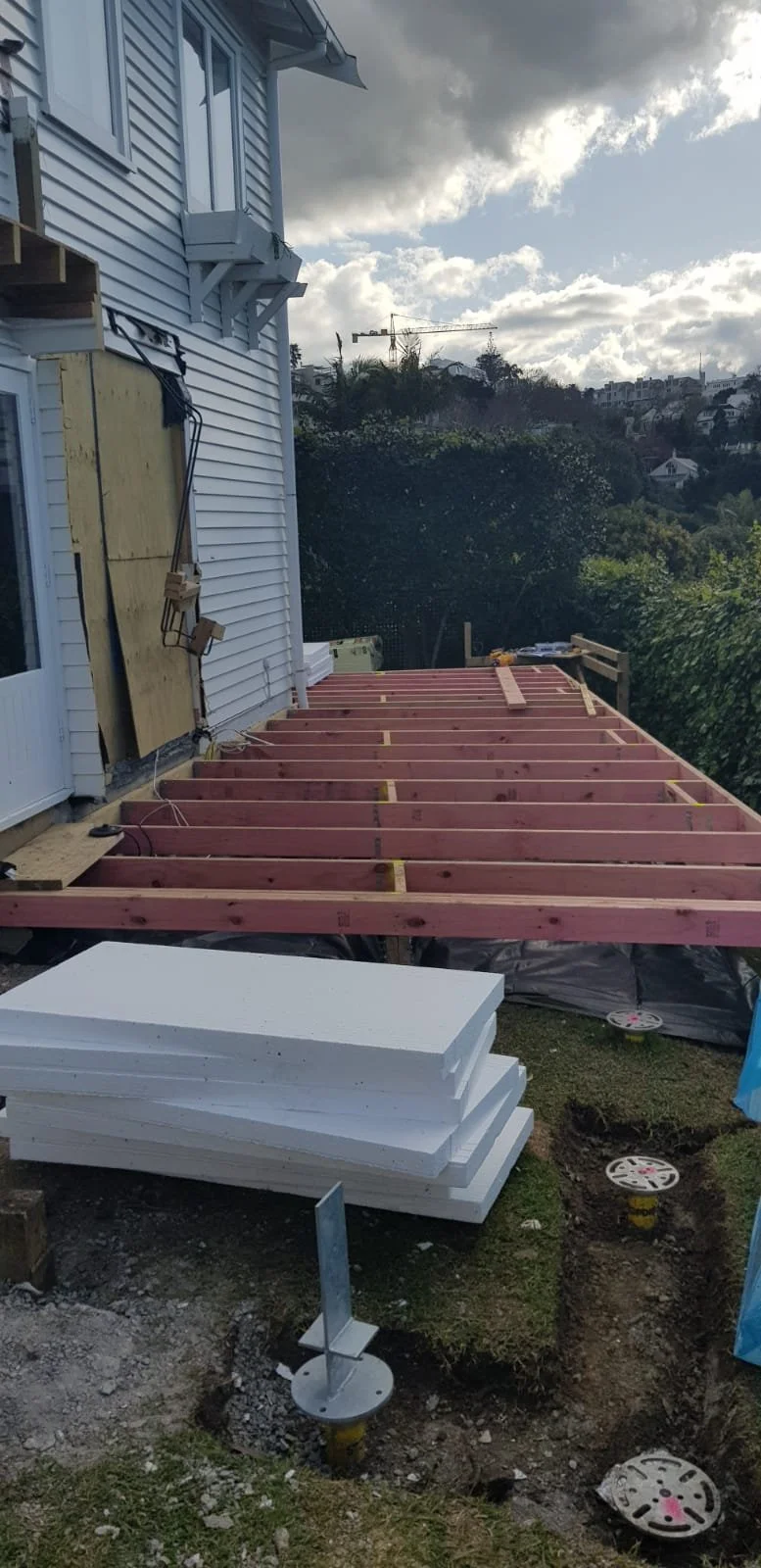 Construction site of an outdoor deck attached to a house, wooden frame partially built, white foam insulation panels on the ground nearby, cloudy sky overhead.