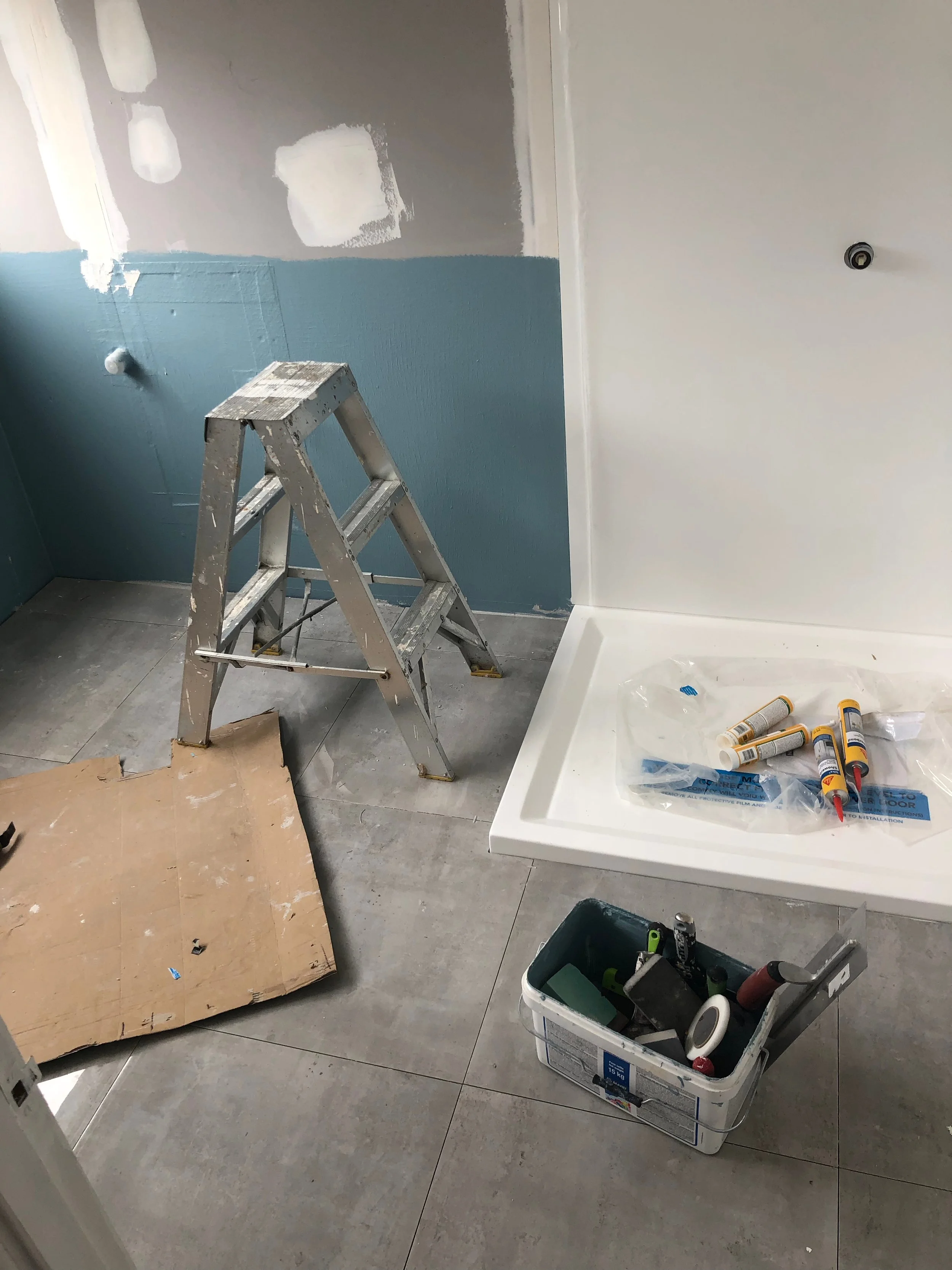A partly renovated bathroom with a ladder, painting supplies, and a shower base on the floor.