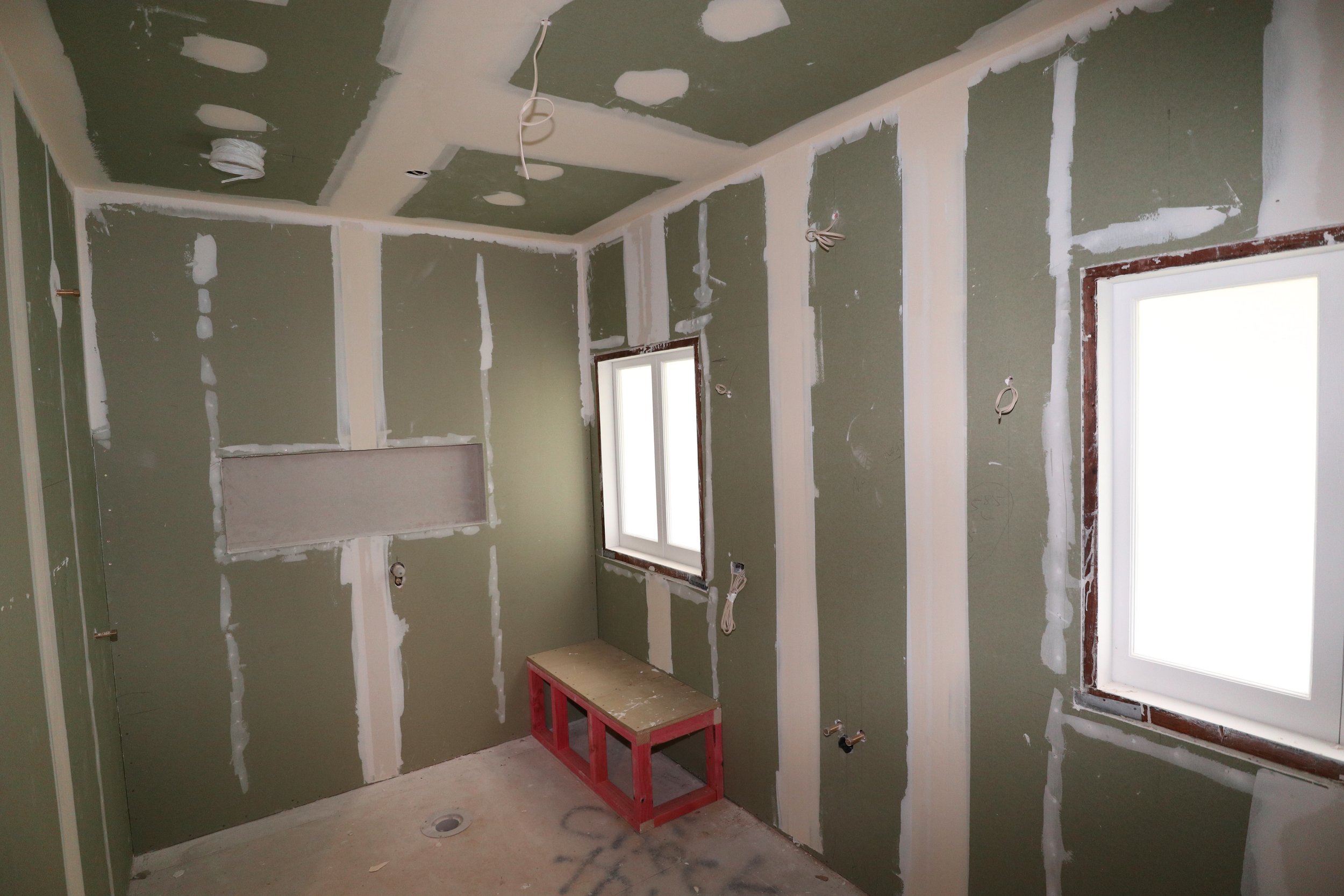 Interior of a room under construction with green drywall, two windows, scattered wiring, and a small red wooden bench on the floor.